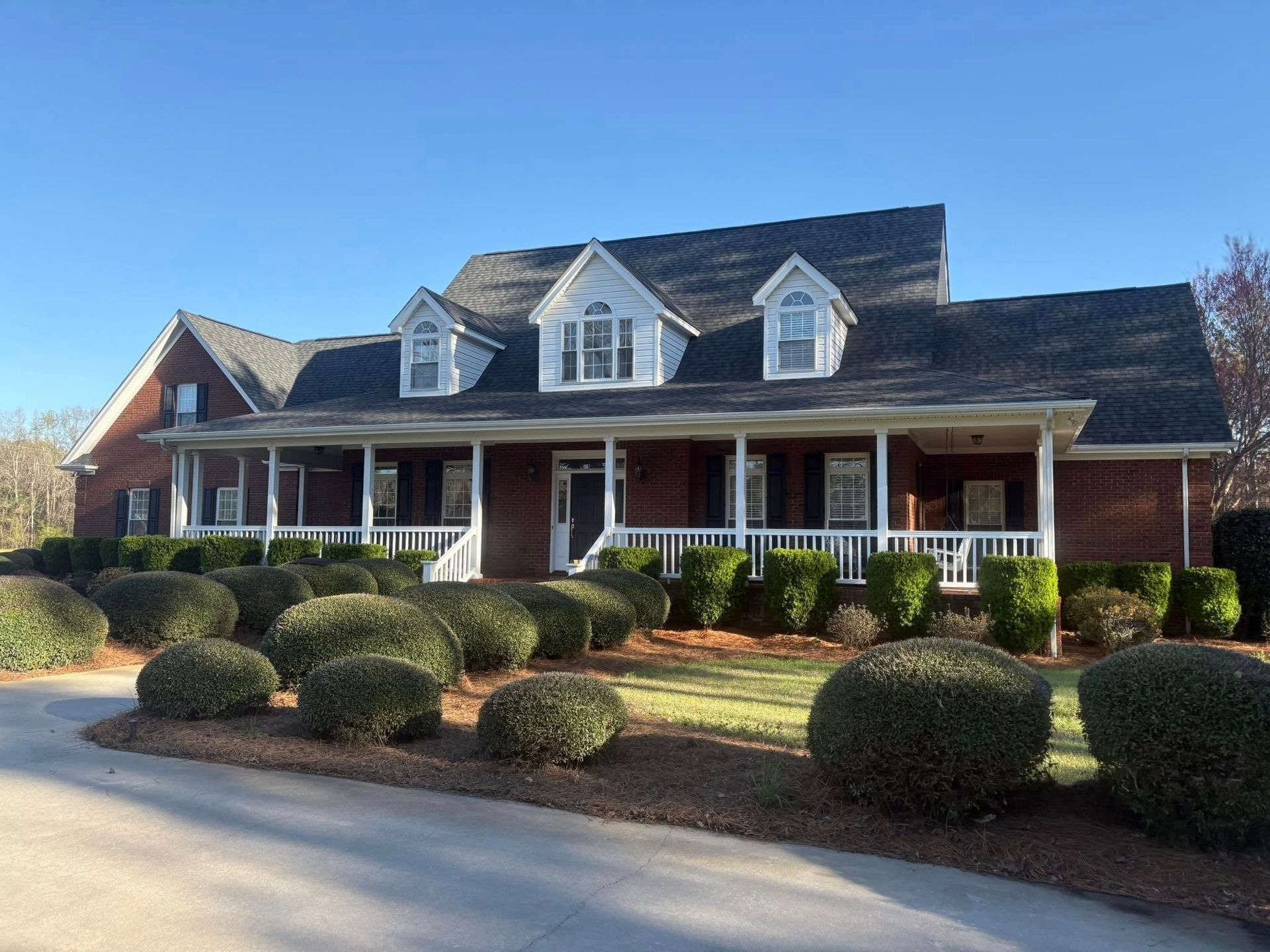 Brown house with a porch, dormers, and rounded bushes in front. Clear, sunny day.