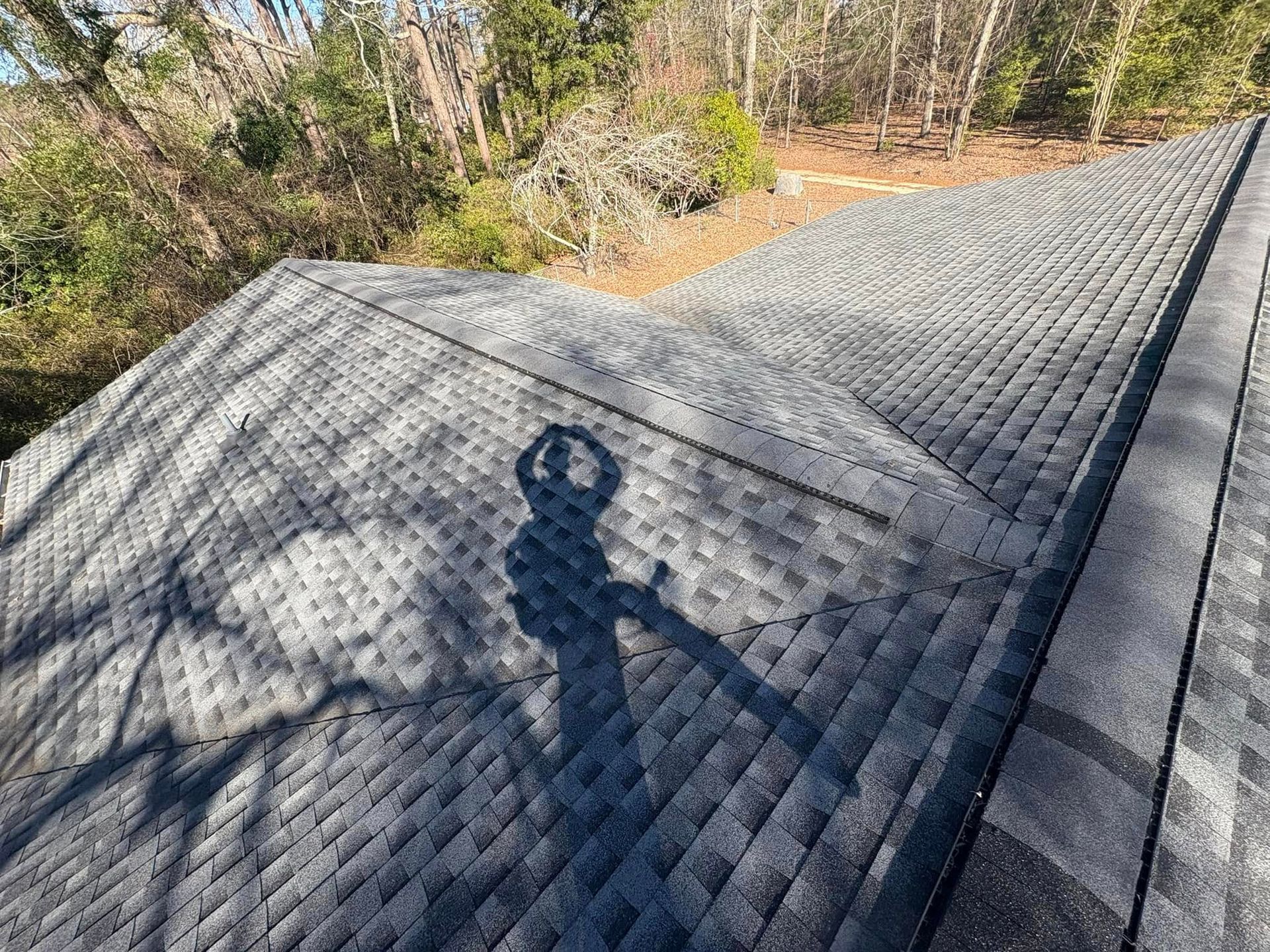 Shadow of a person on a gray shingled roof with trees in the background on a sunny day.