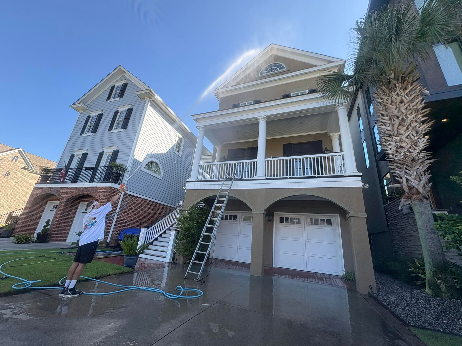 Person spraying water on two-story houses. One house is blue, the other tan, with garages below. Sunny day.