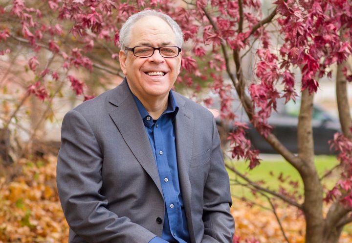 Peter Demetri in a suit and glasses is sitting under a tree with pink flowers.