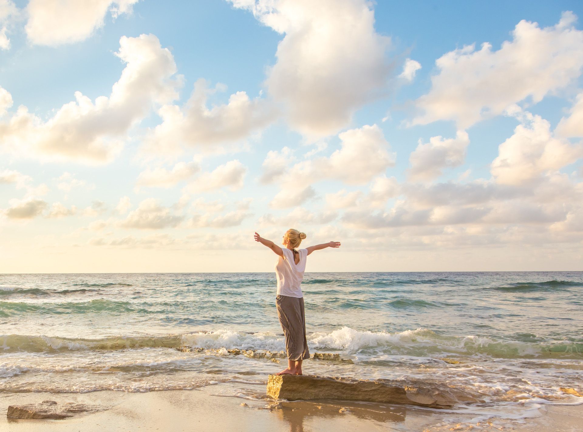 A woman is standing on a rock on the beach with her arms outstretched.