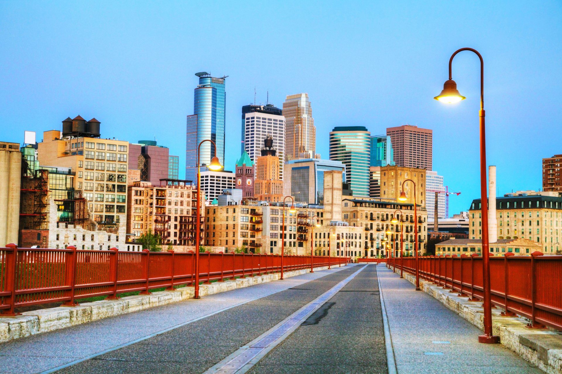 Urban bridge leading to a colorful city skyline at sunset, with red railings and a glowing streetlamp