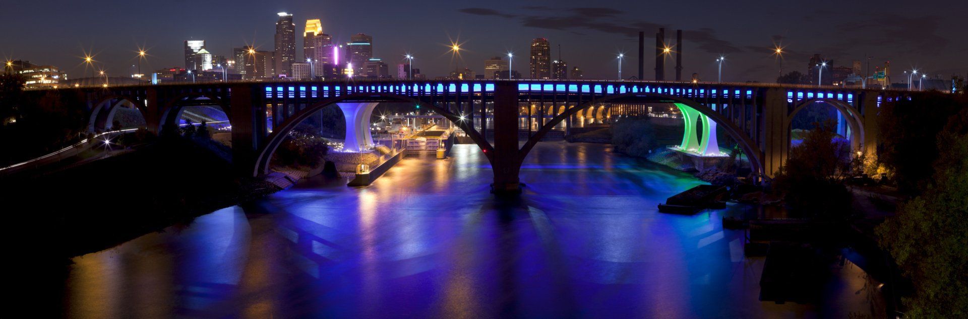 Nighttime city bridge lit blue and purple, reflecting over a river.