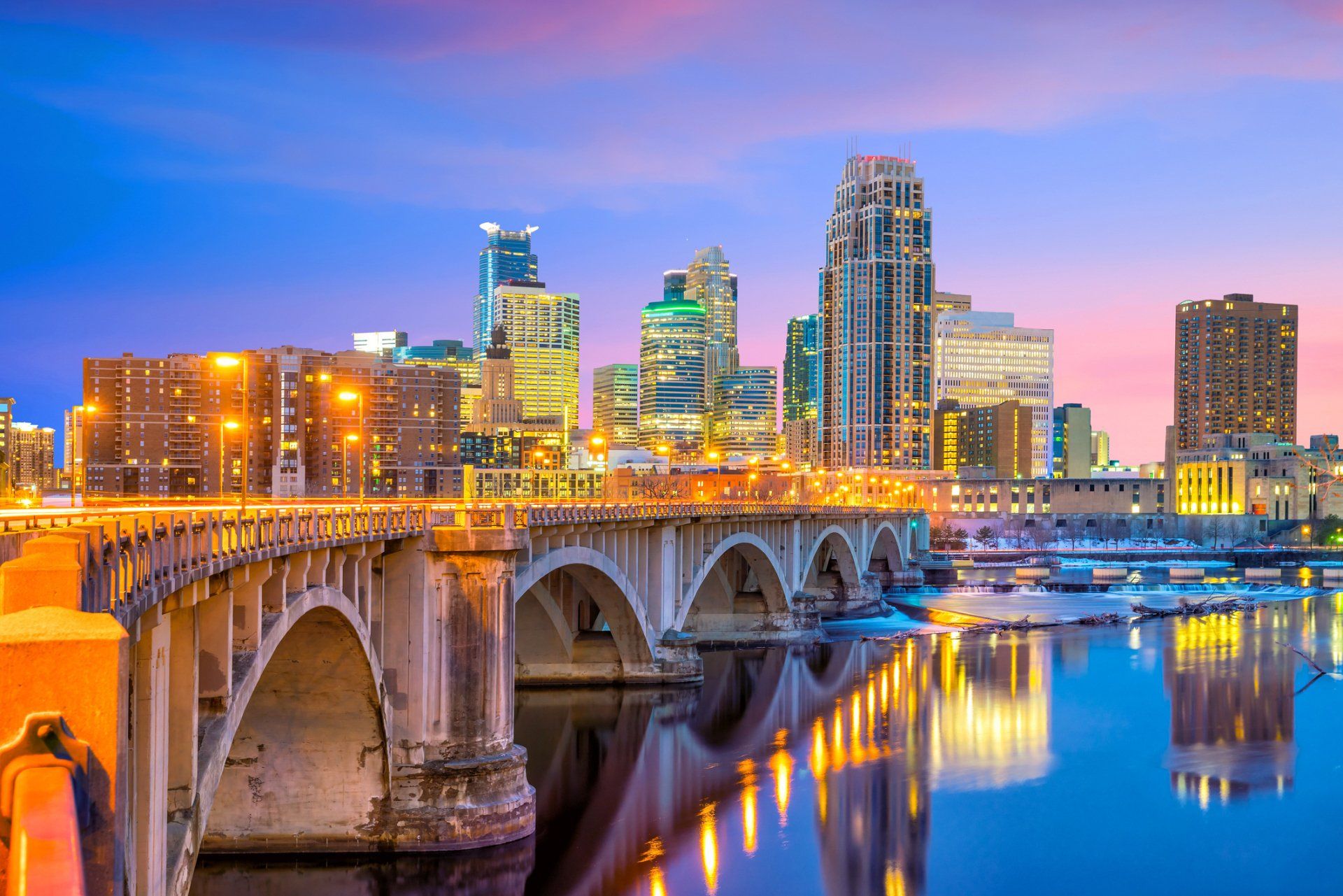 Stone bridge over a river with a lit city skyline at sunset