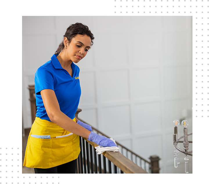 Woman in blue polo and yellow apron cleaning a railing with gloves in a bright room