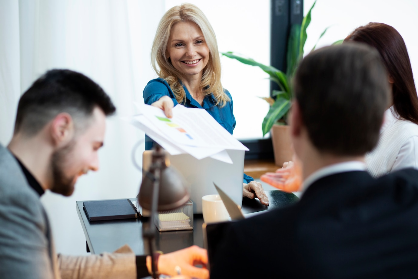A woman is holding a piece of paper in front of a group of people sitting around a table.