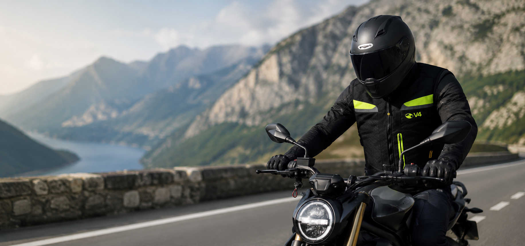 Motociclista in tenuta nera che percorre una strada di montagna costeggiando un lago.