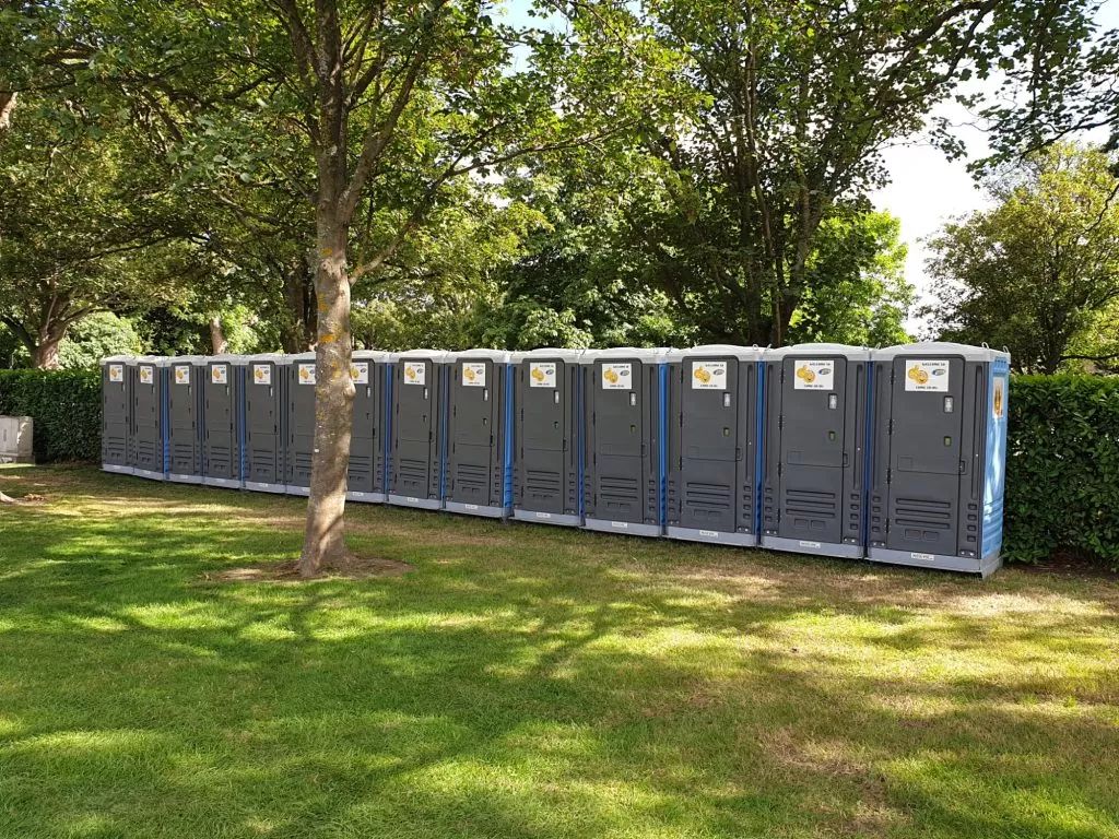 Row of portable toilets on a grassy lawn, near trees and bushes.
