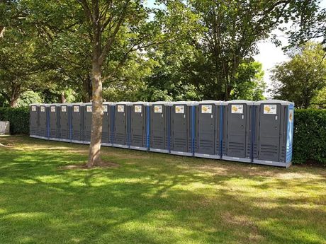 Row of portable toilets on green grass, next to trees and bushes.