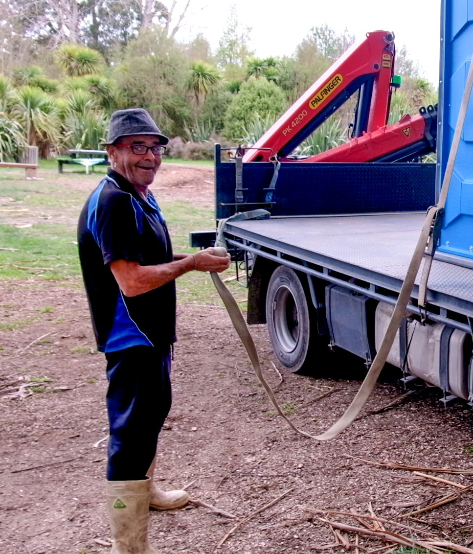 Man holding strap next to truck with a red crane and portable toilet on flatbed. Outdoors.