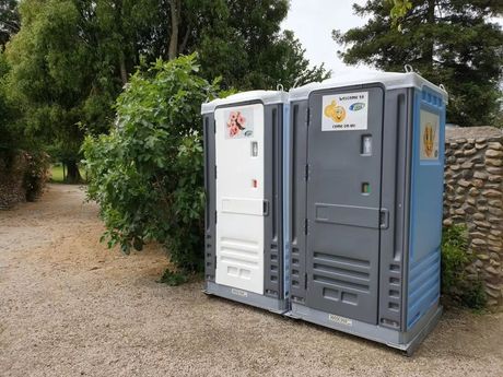 Two portable toilets, white and grey-blue, sit on gravel beside a path with a leafy backdrop.