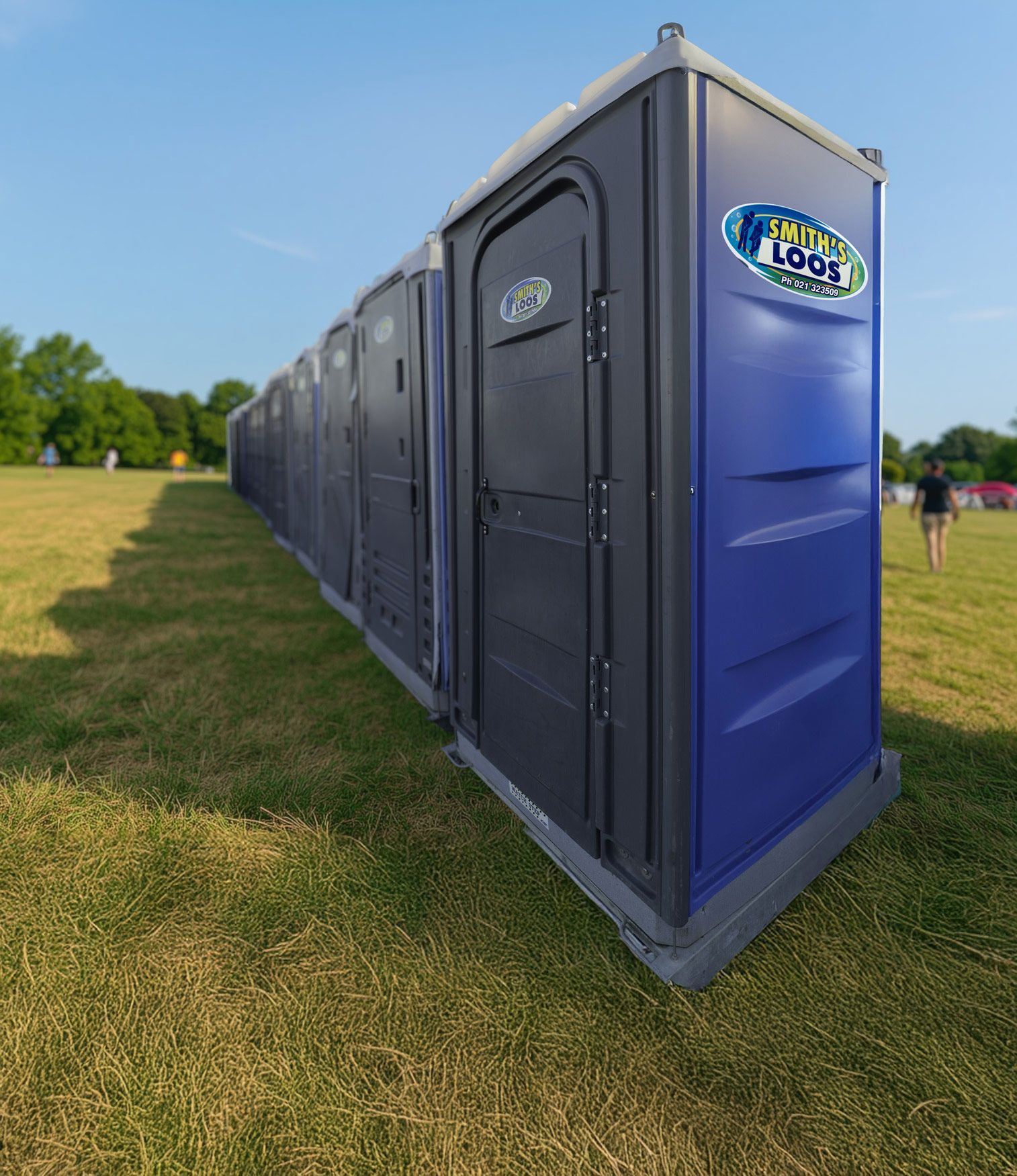 Row of blue and black portable toilets on a grassy field; a person walks in the distance.