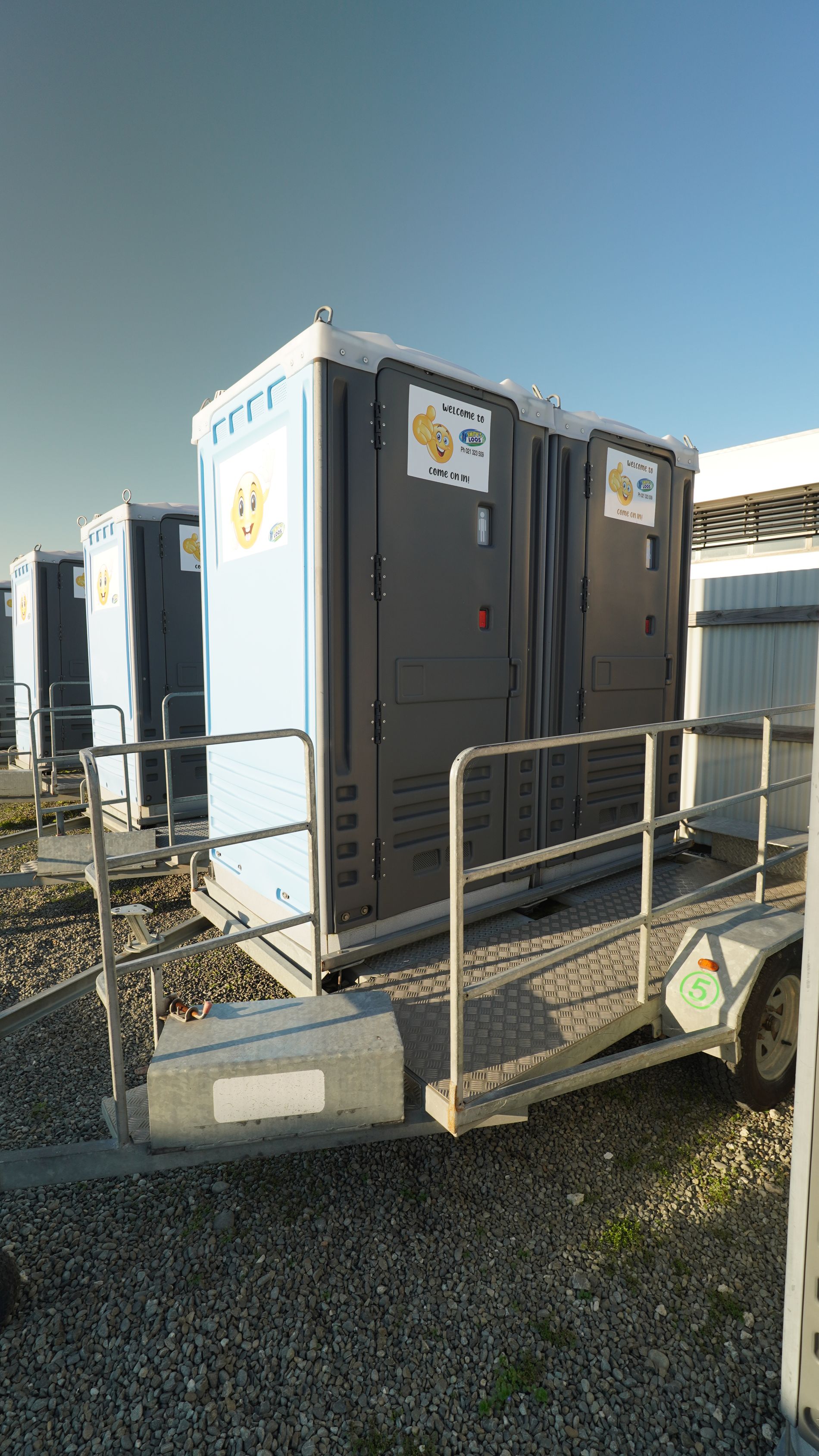 Portable toilets on a trailer, outside. Light blue and dark grey, with a ramp.