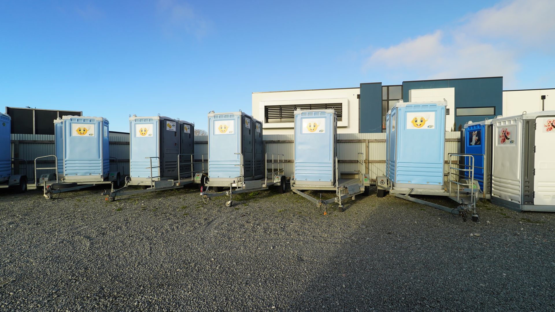 Several blue portable toilets on trailers parked on gravel, with a building in the background.