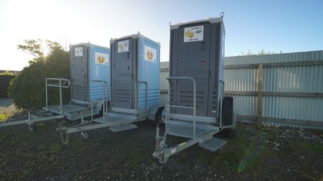 Three portable toilets on trailers, parked outdoors. Gray and blue units with ramps, near a fence.