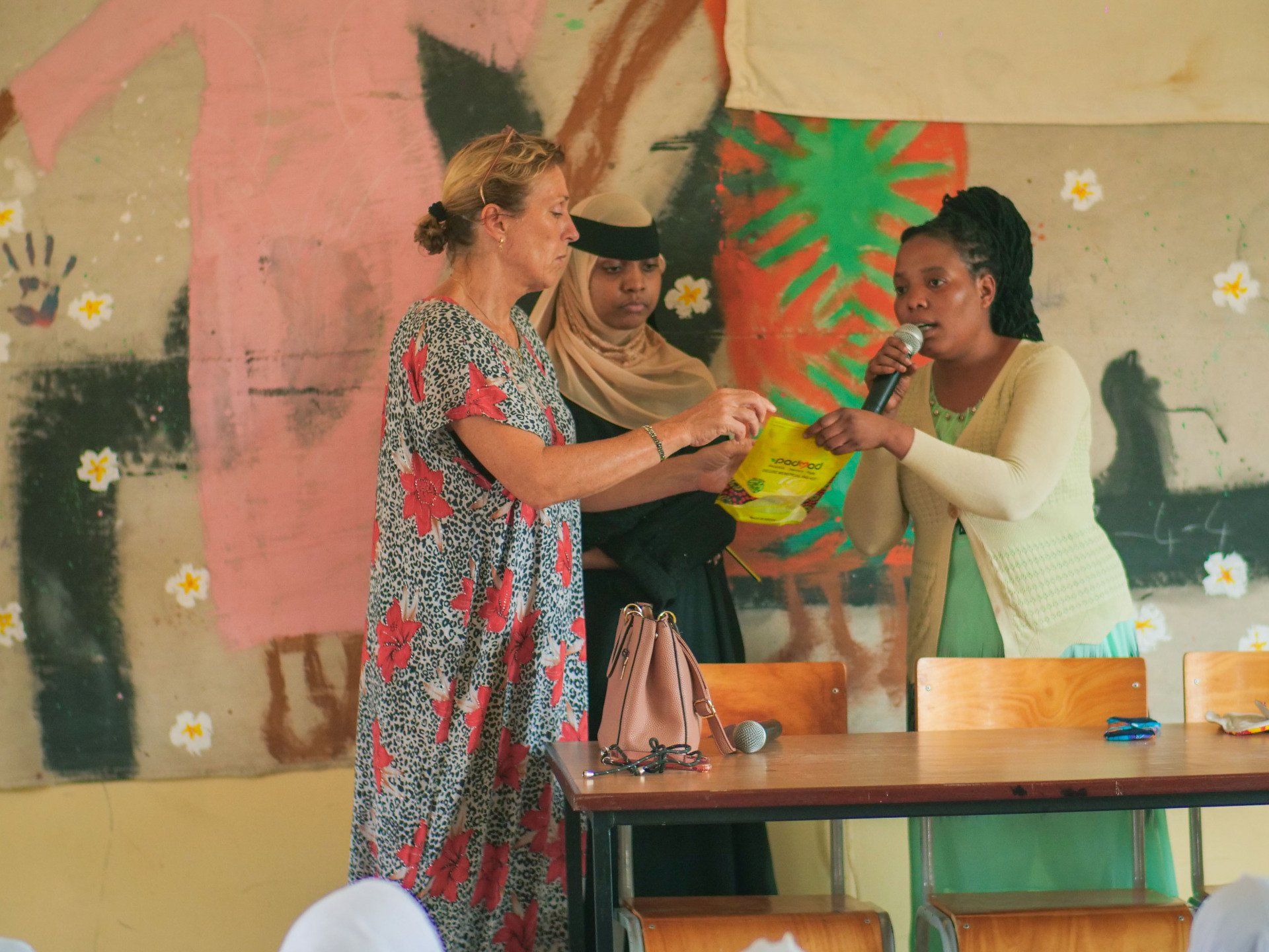 “Community gathering in Shela where participants exchange items while one woman speaks into a microphone during a local event