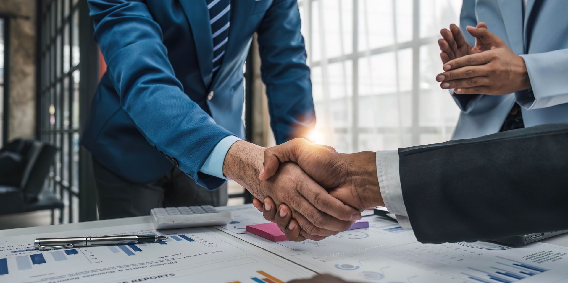 Two professionals in business suits shake hands over a table covered with financial charts and documents.