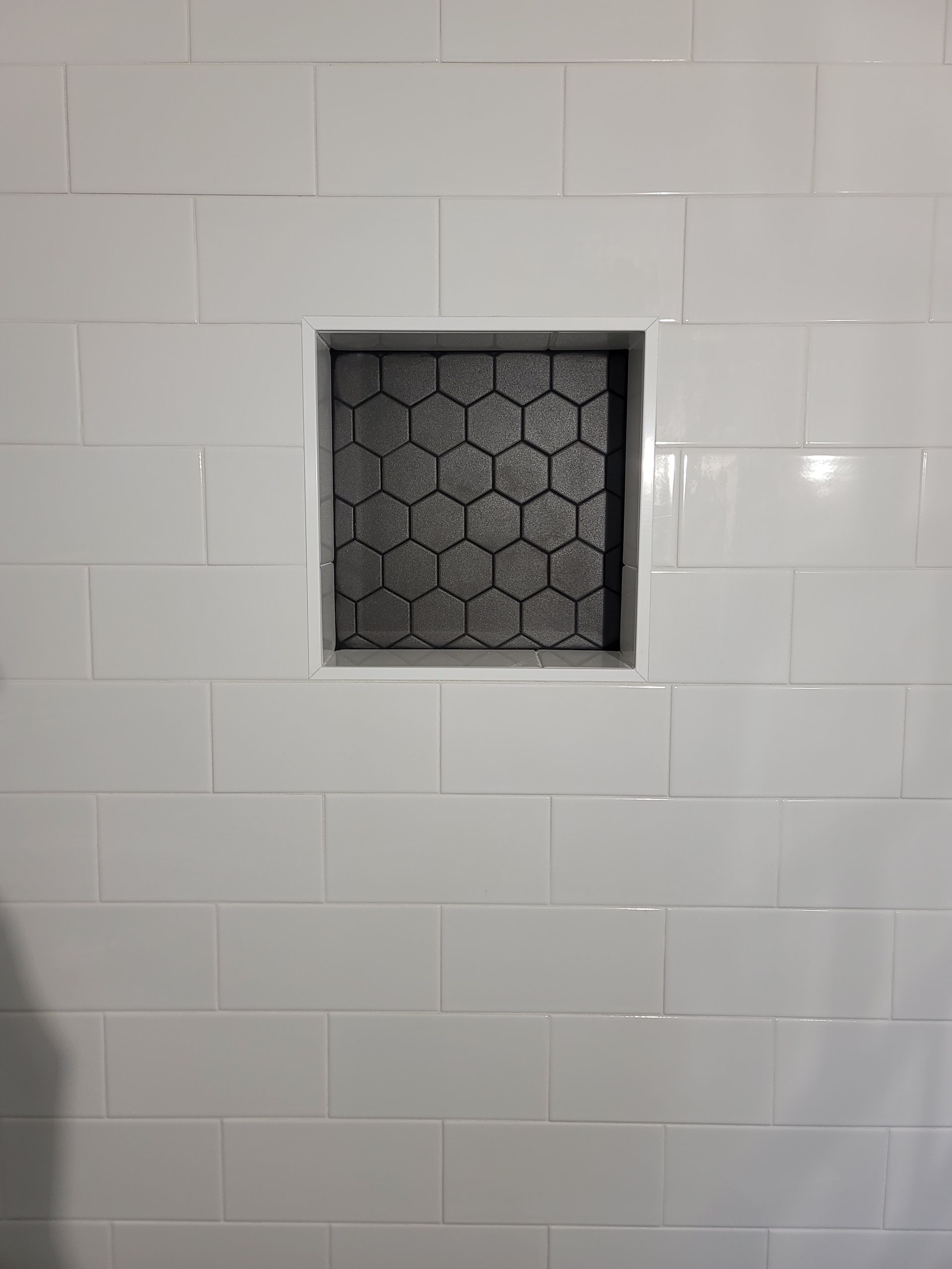 White tiled wall with a small square recessed vent or niche covered by hexagonal mesh.