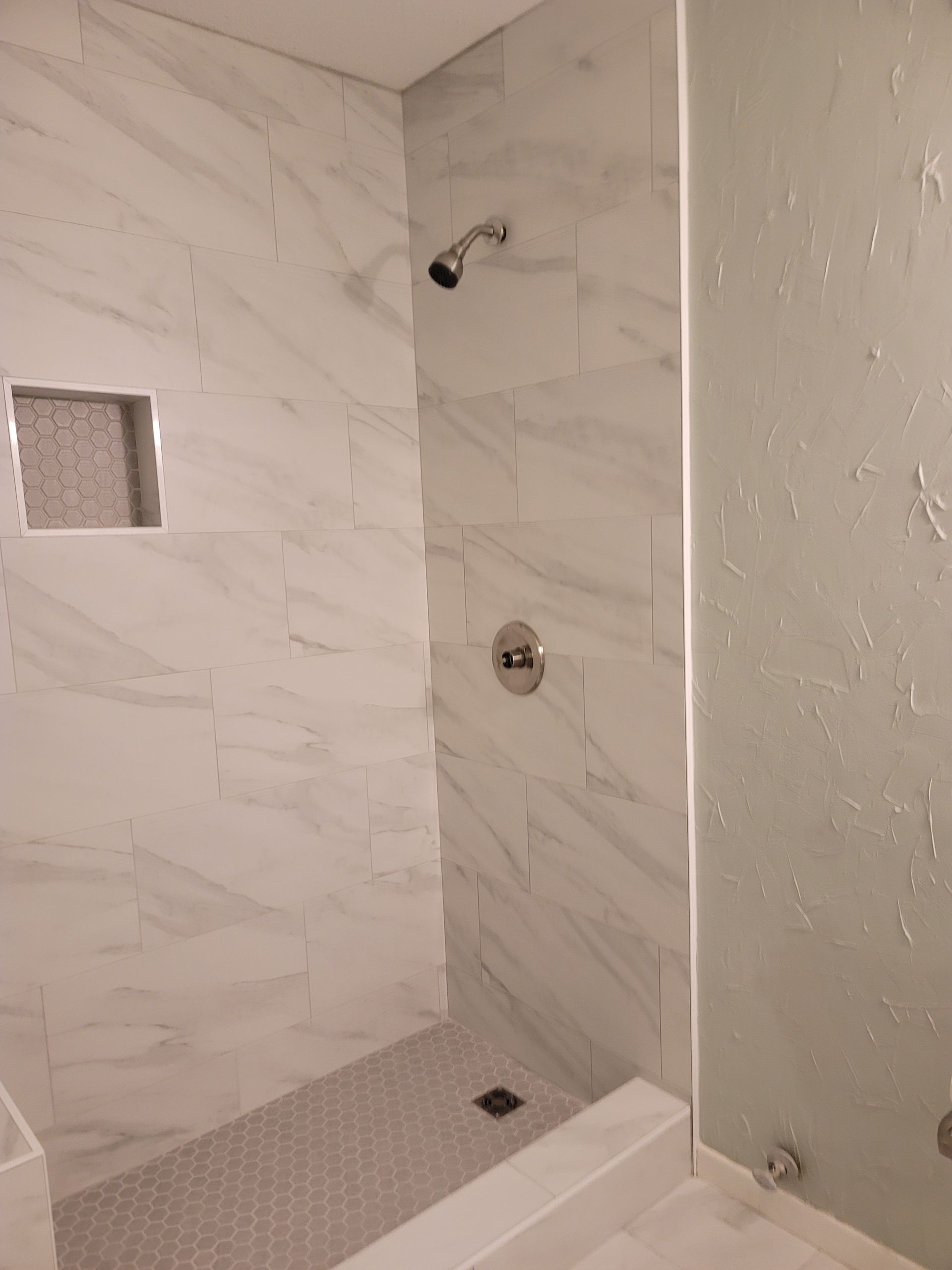 Empty marble-tiled shower with two wall fixtures, a square niche, and a glass partition.