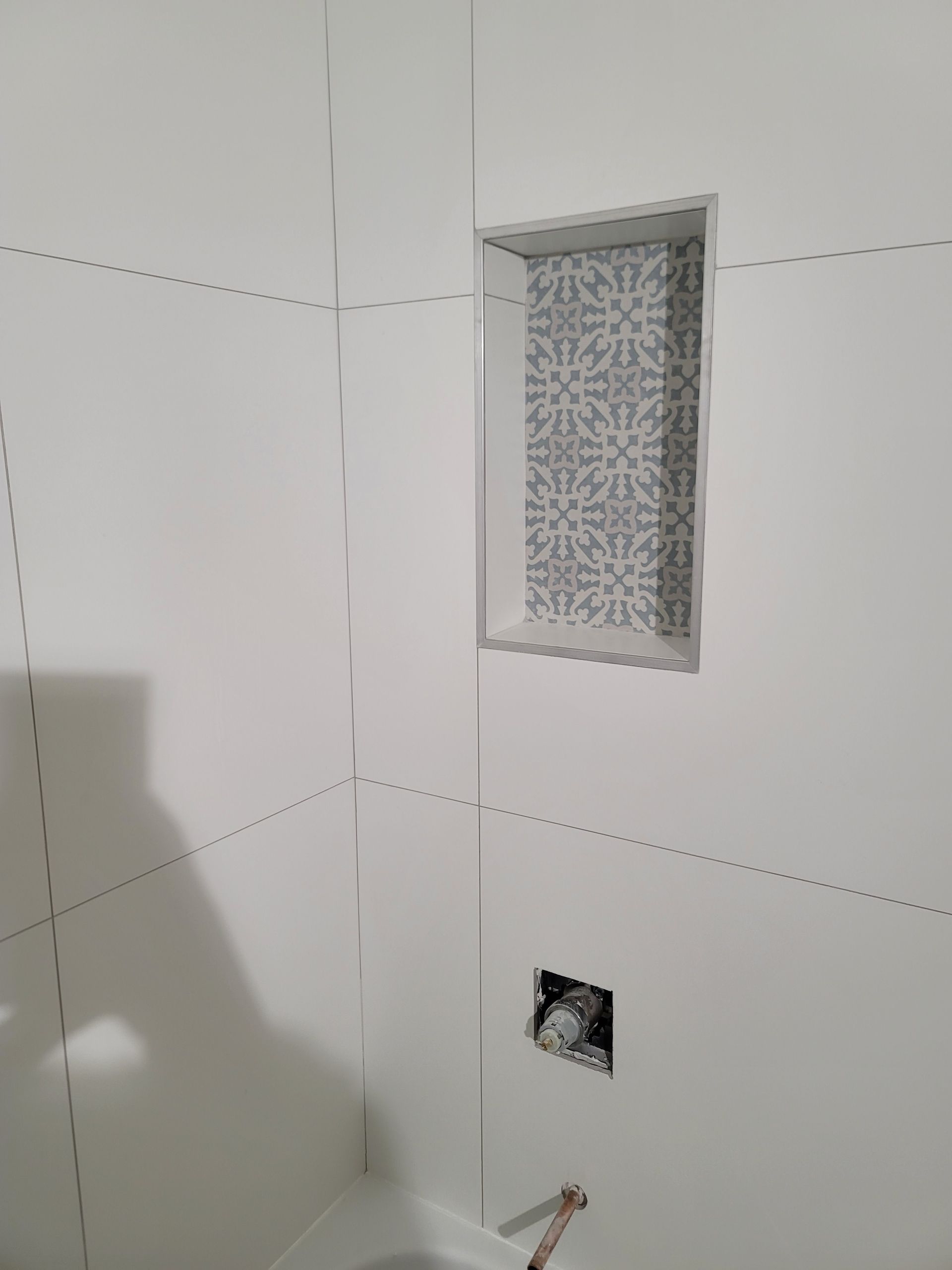 Tiled shower wall with a small recessed niche, exposed pipe opening, and unfinished plumbing hole near the floor