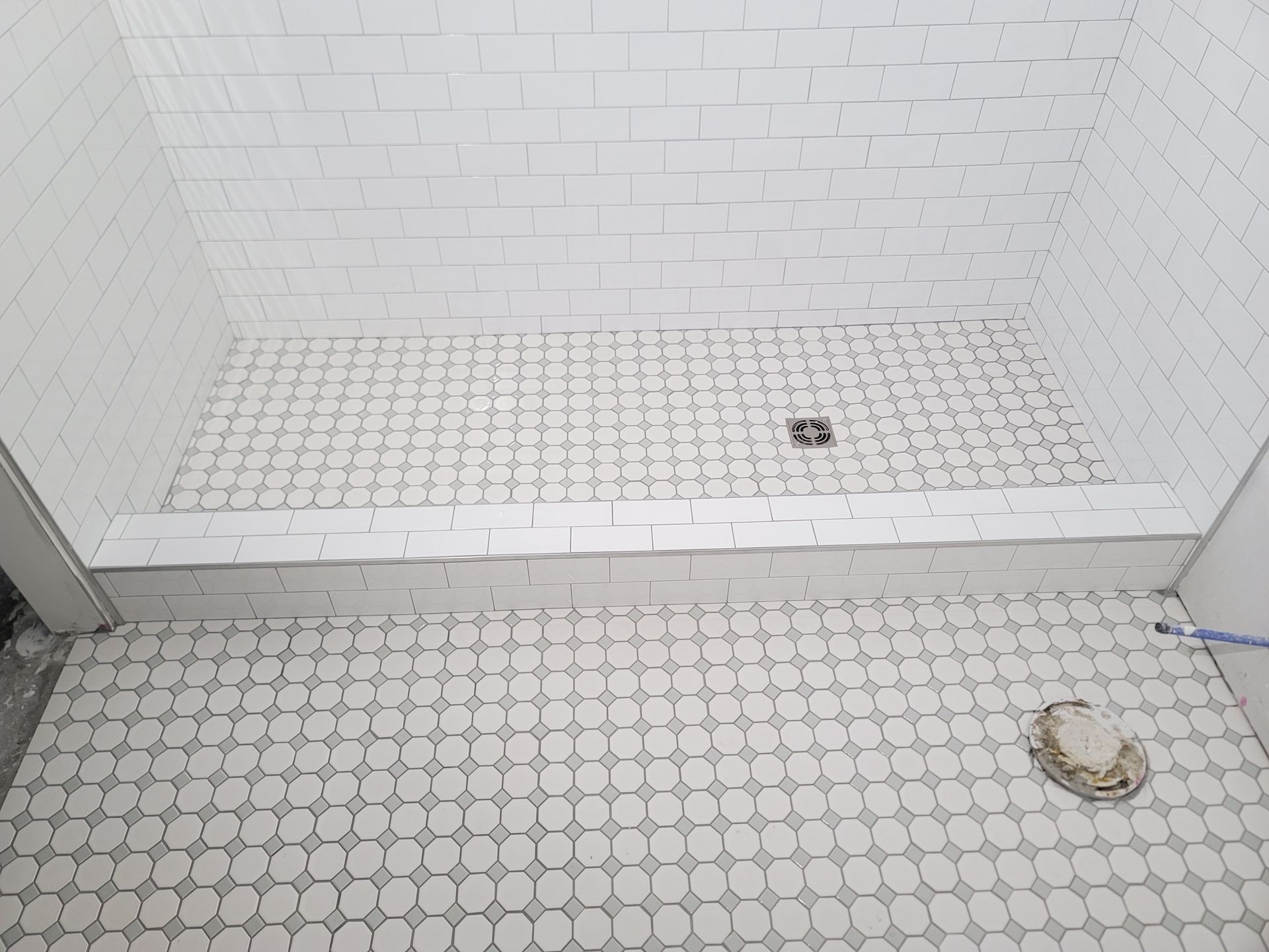 White tiled shower stall with a bench, floor drain, and handheld shower head on the floor