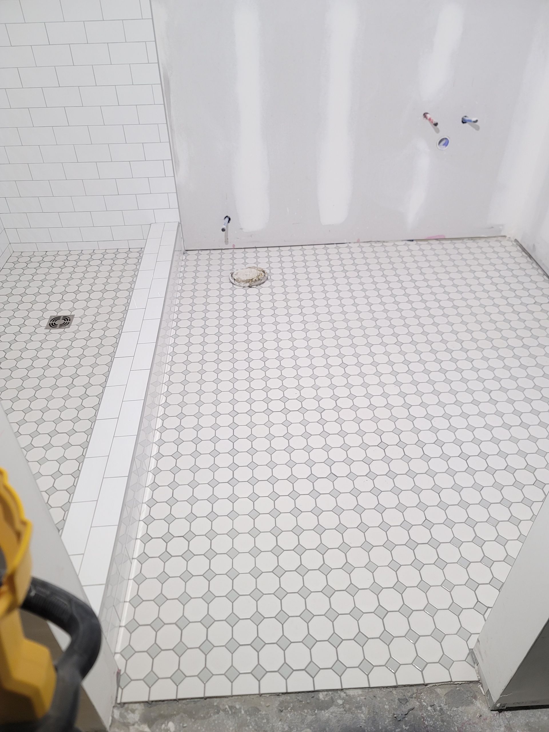 Partially tiled bathroom floor and shower area with white walls and exposed plumbing
