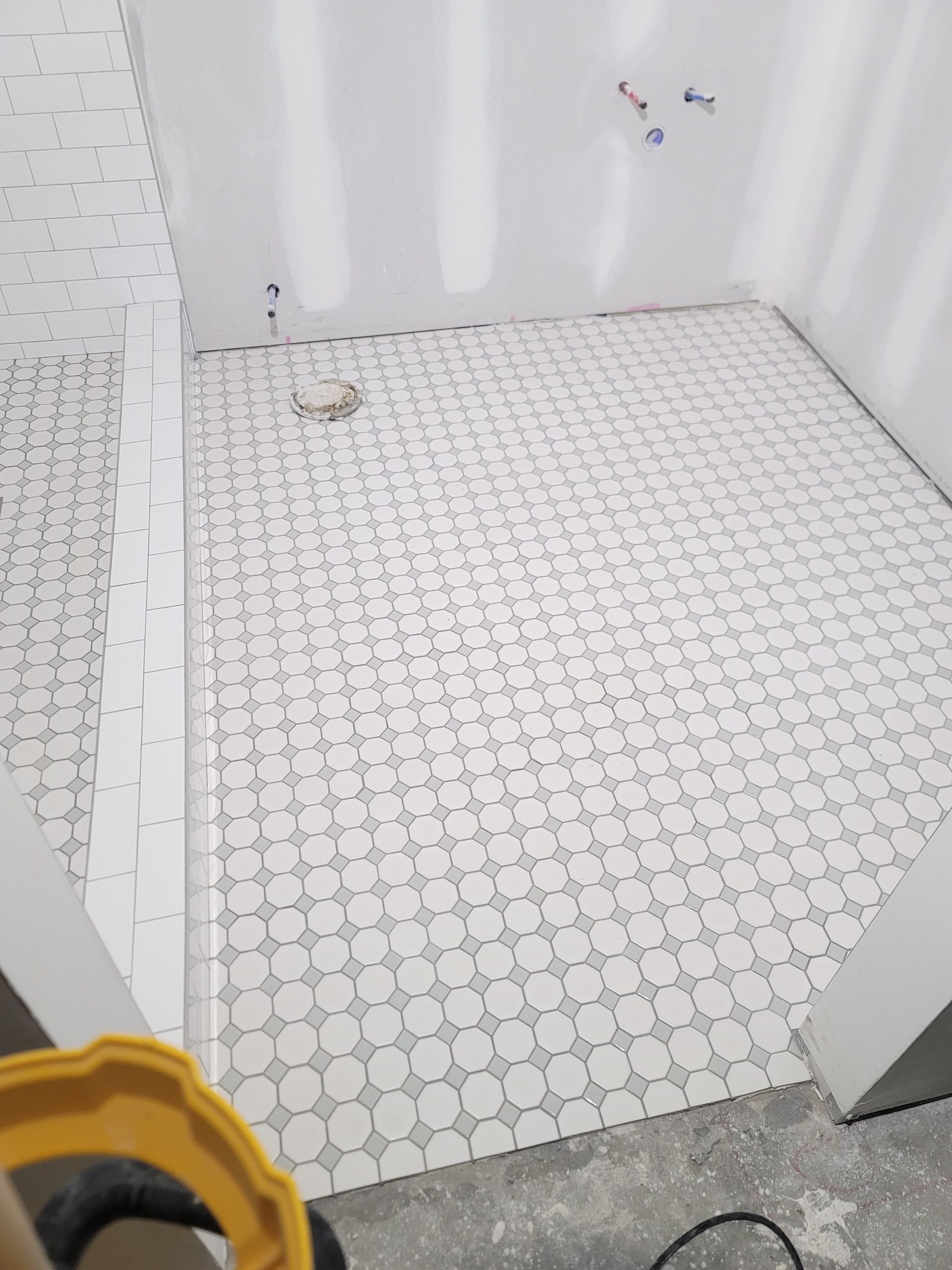 Empty small bathroom with white tiled floor and walls, and a yellow bucket edge in the foreground
