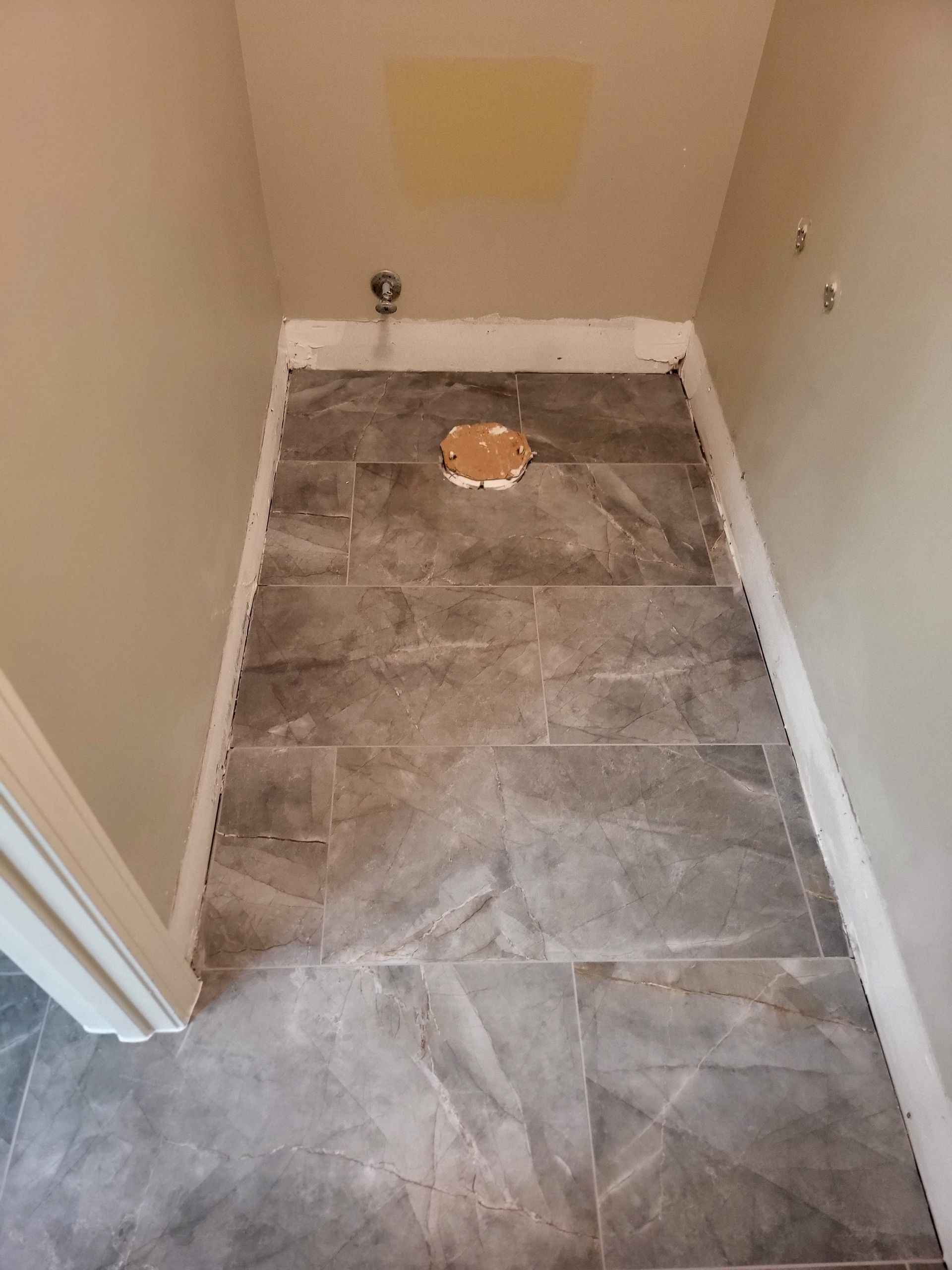 Narrow tiled hallway or closet floor with a drain, orange cap, and a pale patch on the far wall.