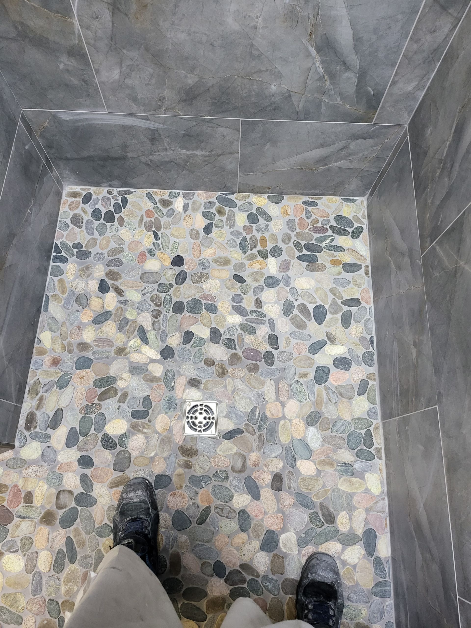 Top-down view of a pebble-floored shower with dark tiled walls and a drain, feet visible at the bottom.