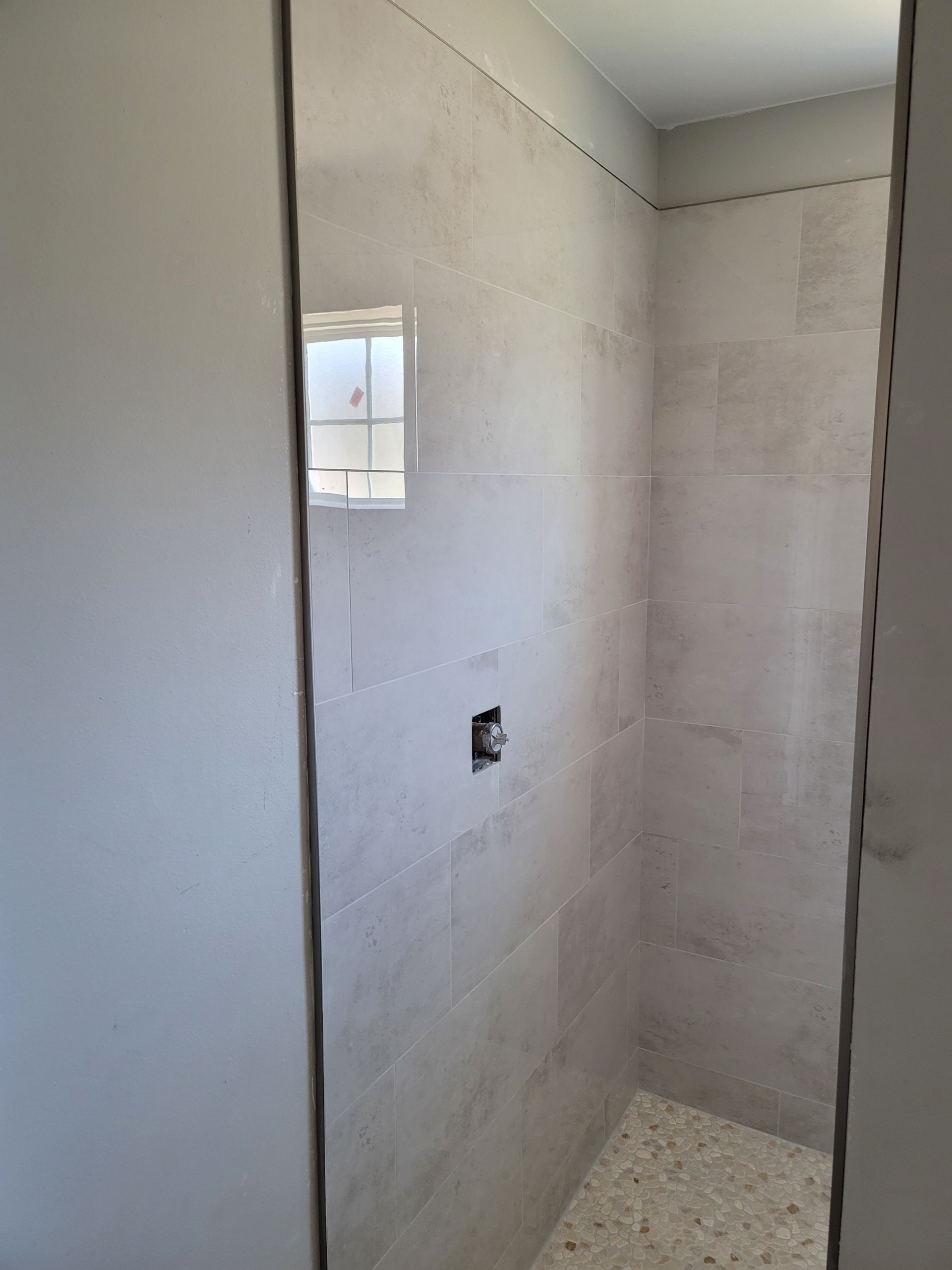 Narrow unfinished tiled shower stall with a small window and exposed plumbing opening