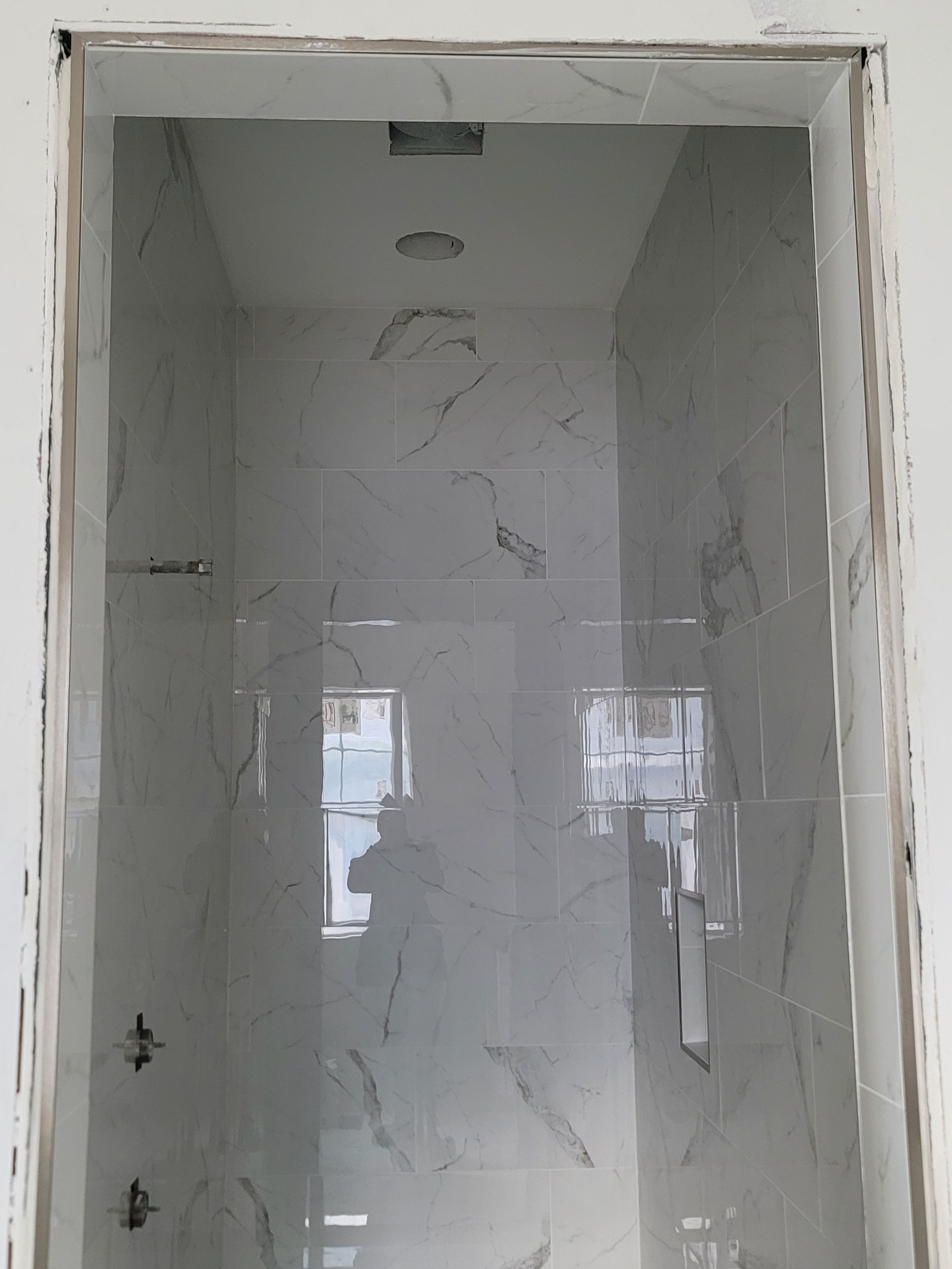 Narrow tiled shower stall with white walls and a recessed ceiling light, viewed through a doorway