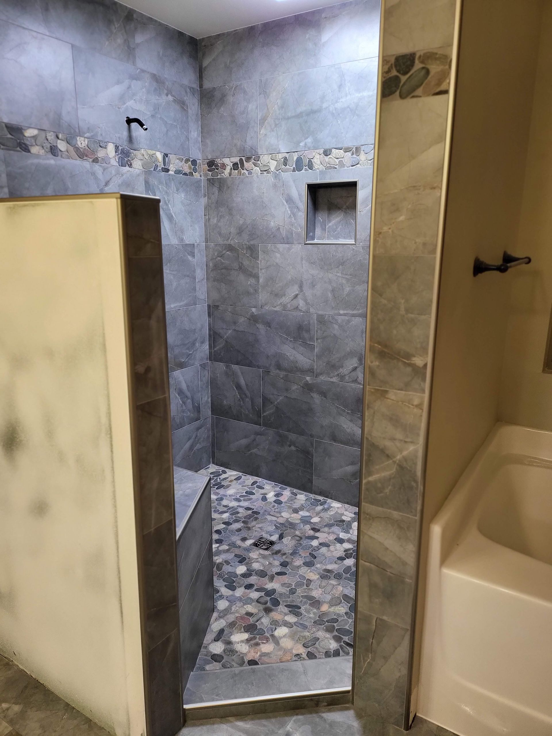 Partially tiled shower with gray stone walls, pebble floor, and a bathtub beside it