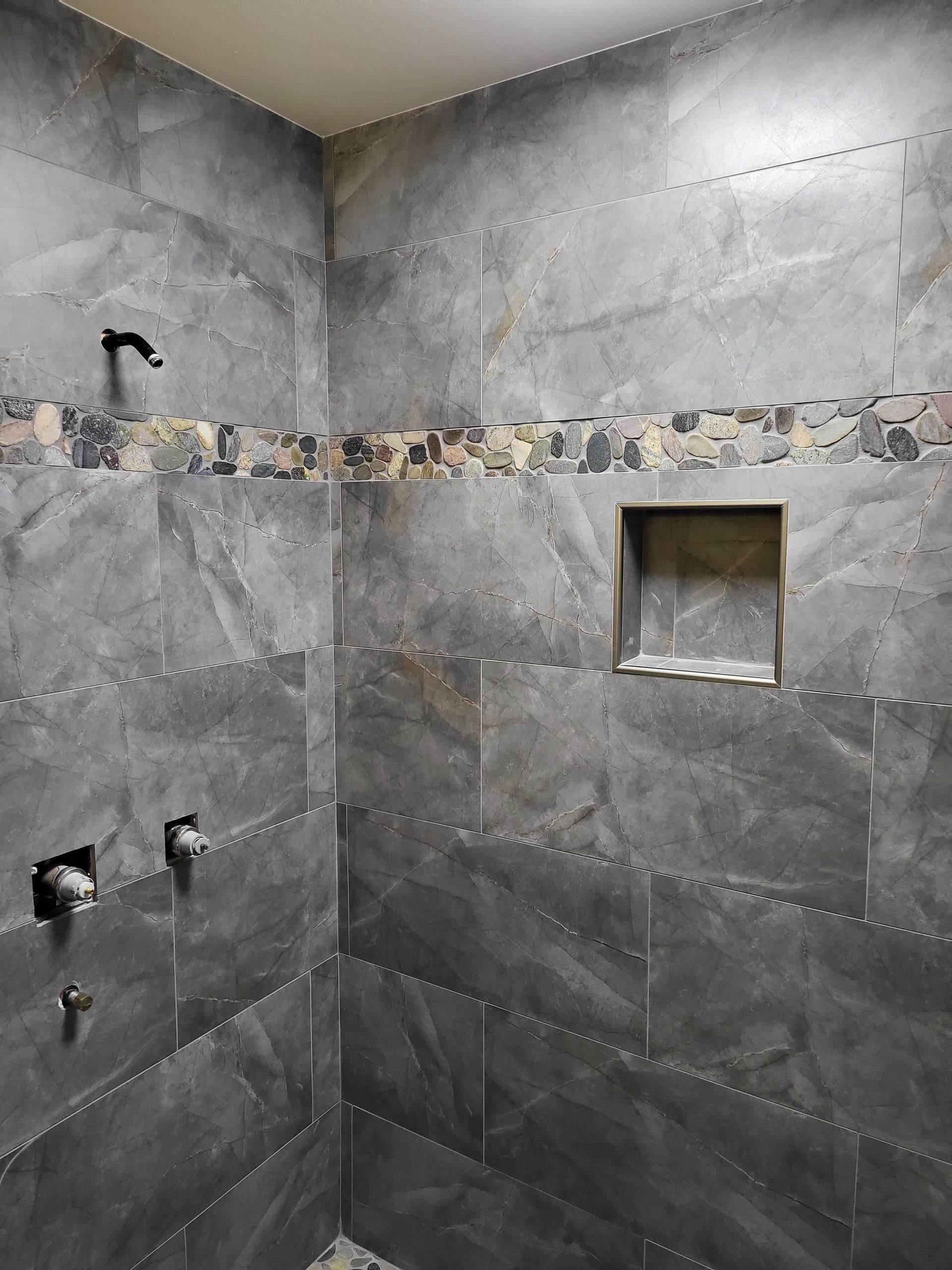 Tiled shower stall with gray stone walls, decorative mosaic border, and recessed niche.