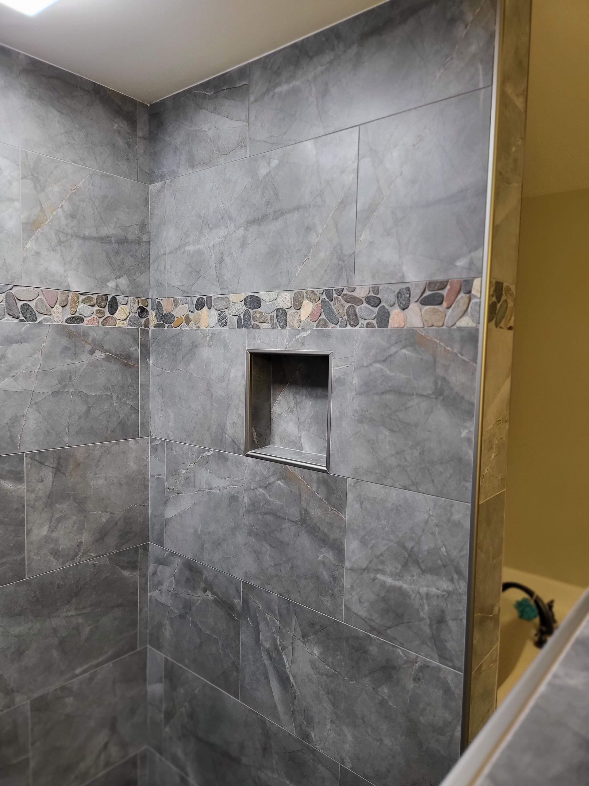 Tiled shower wall with gray stone tiles, decorative mosaic band, and recessed shelf niche