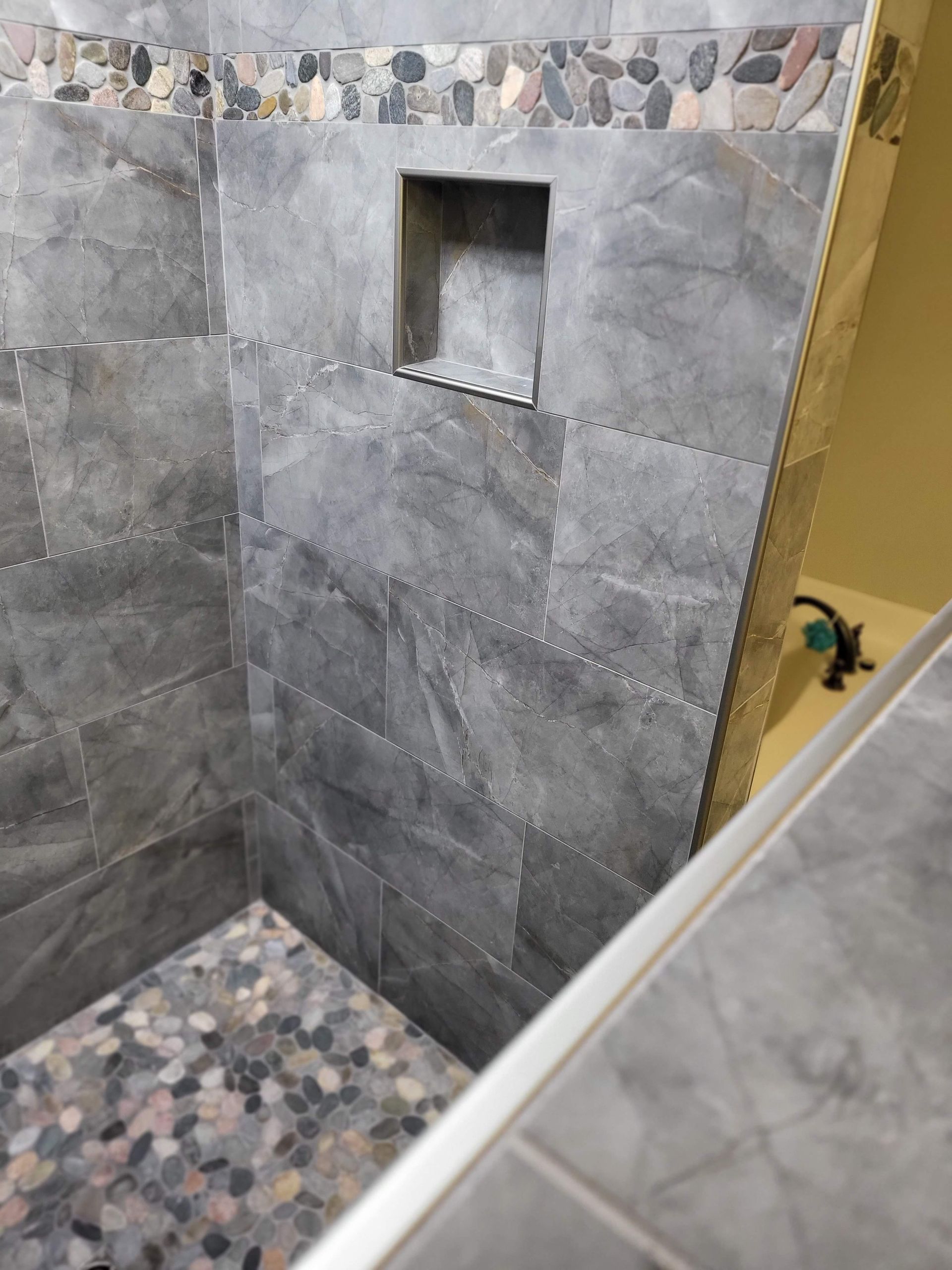 Gray tiled shower with pebble trim and a small recessed shelf niche