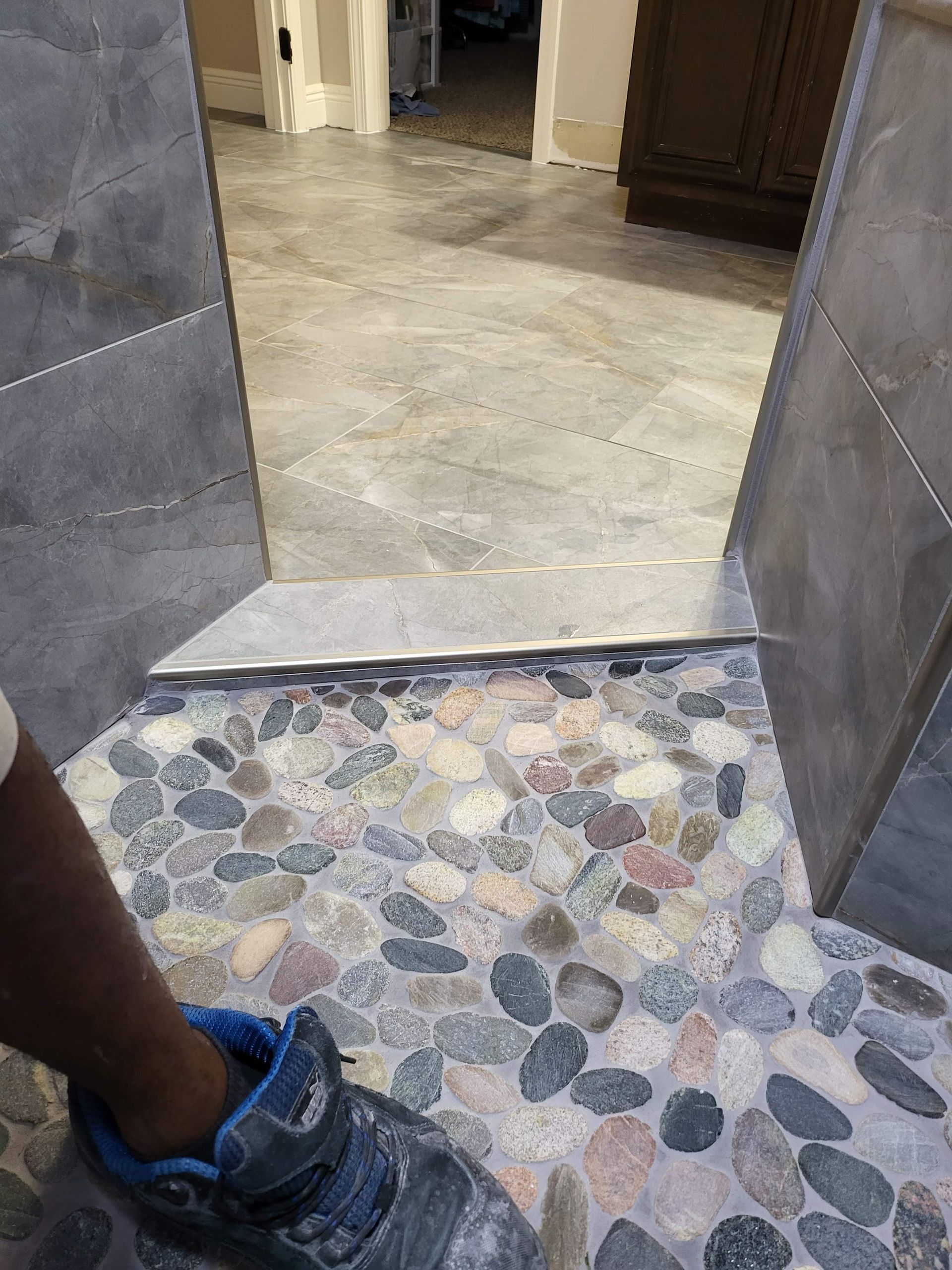 Pebbled floor at a doorway leading into a hallway with beige tile and a dark cabinet.