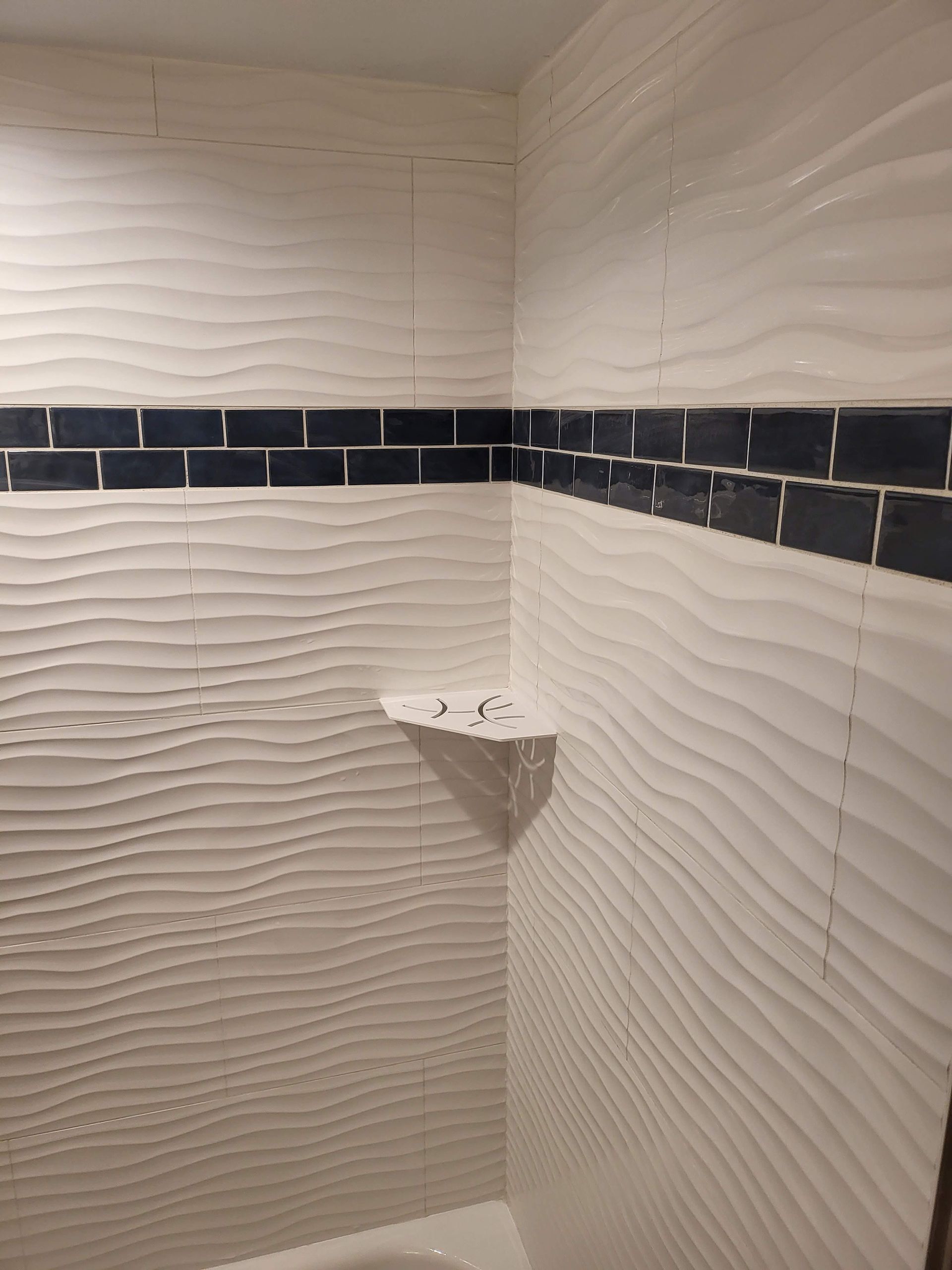 Empty tiled shower corner with dark horizontal accent strip and a small corner shelf