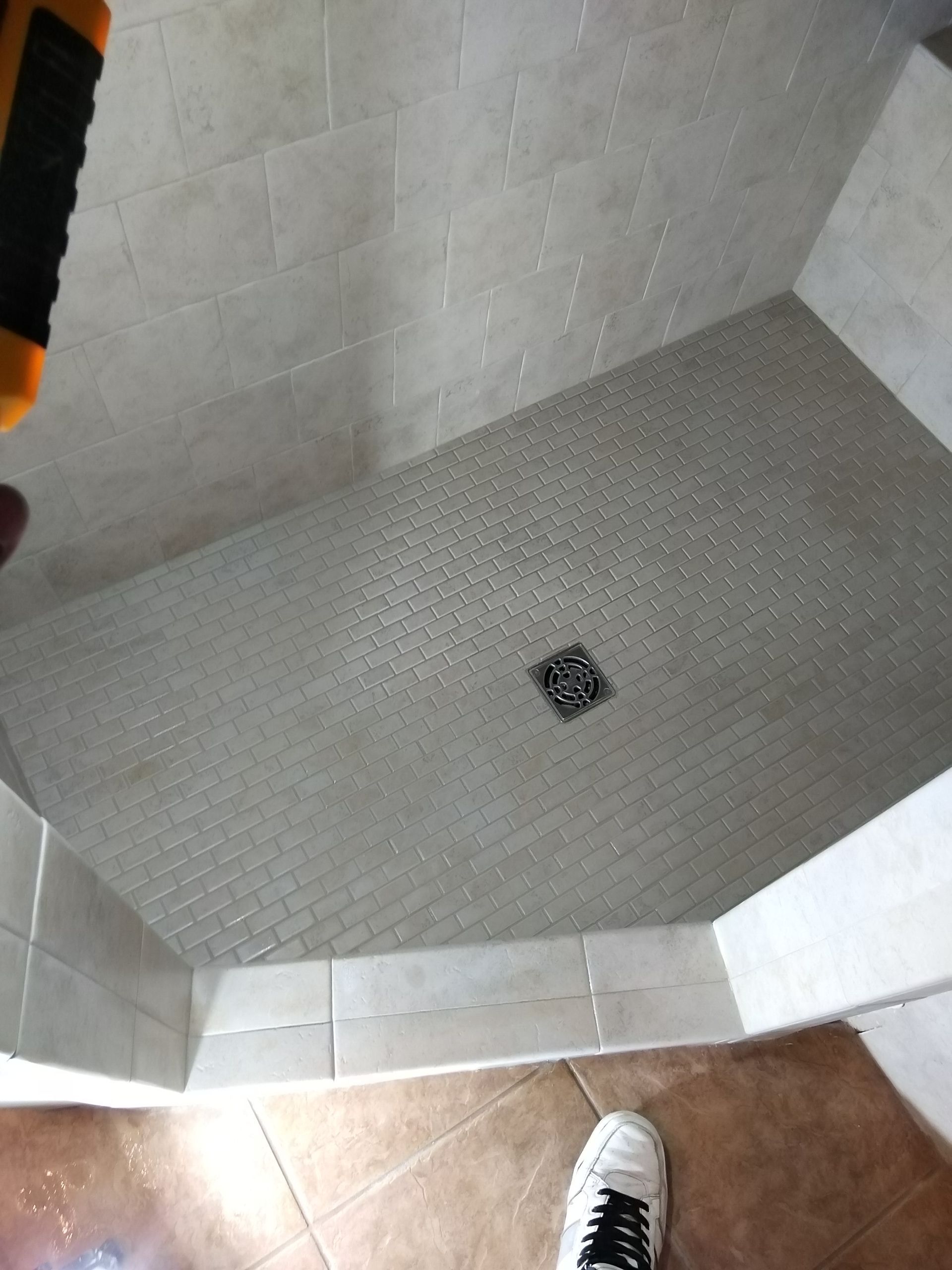 Tiled shower stall with a small drain and visible shoes at the bottom edge