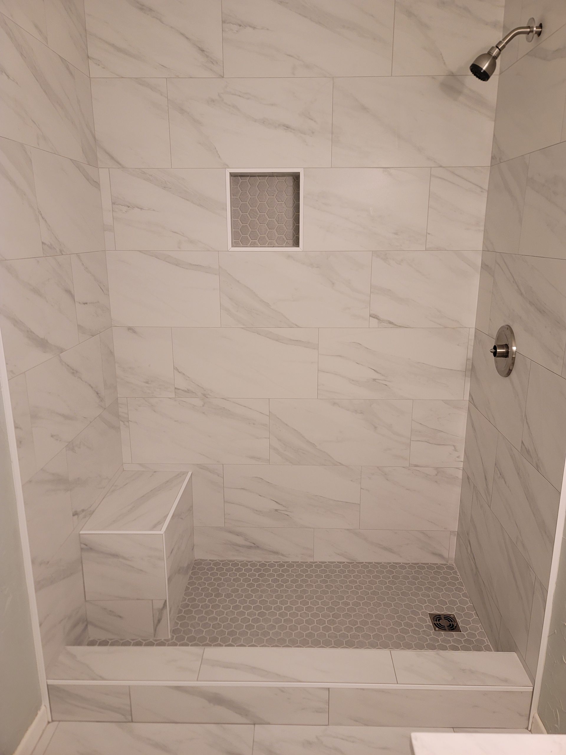 Empty tiled shower with bench, square niche, and drain in a beige marble-patterned bathroom.