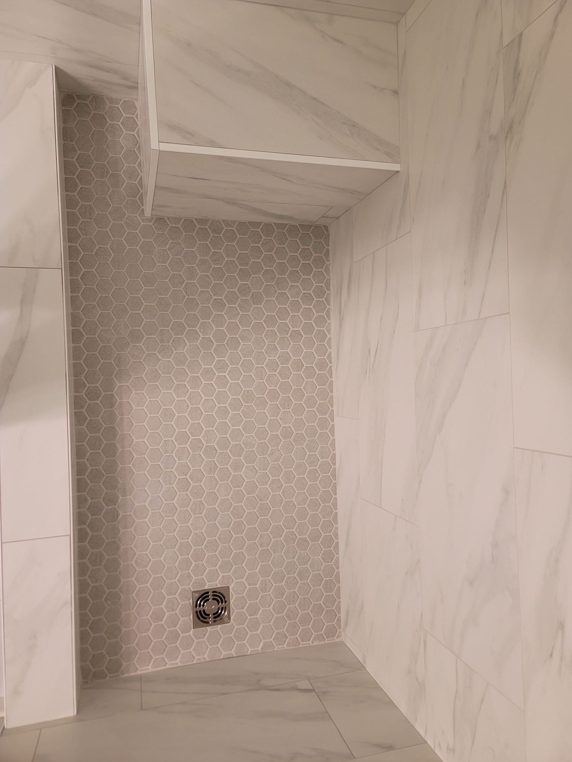 Empty tiled shower nook with a small drain opening and marble-look walls.