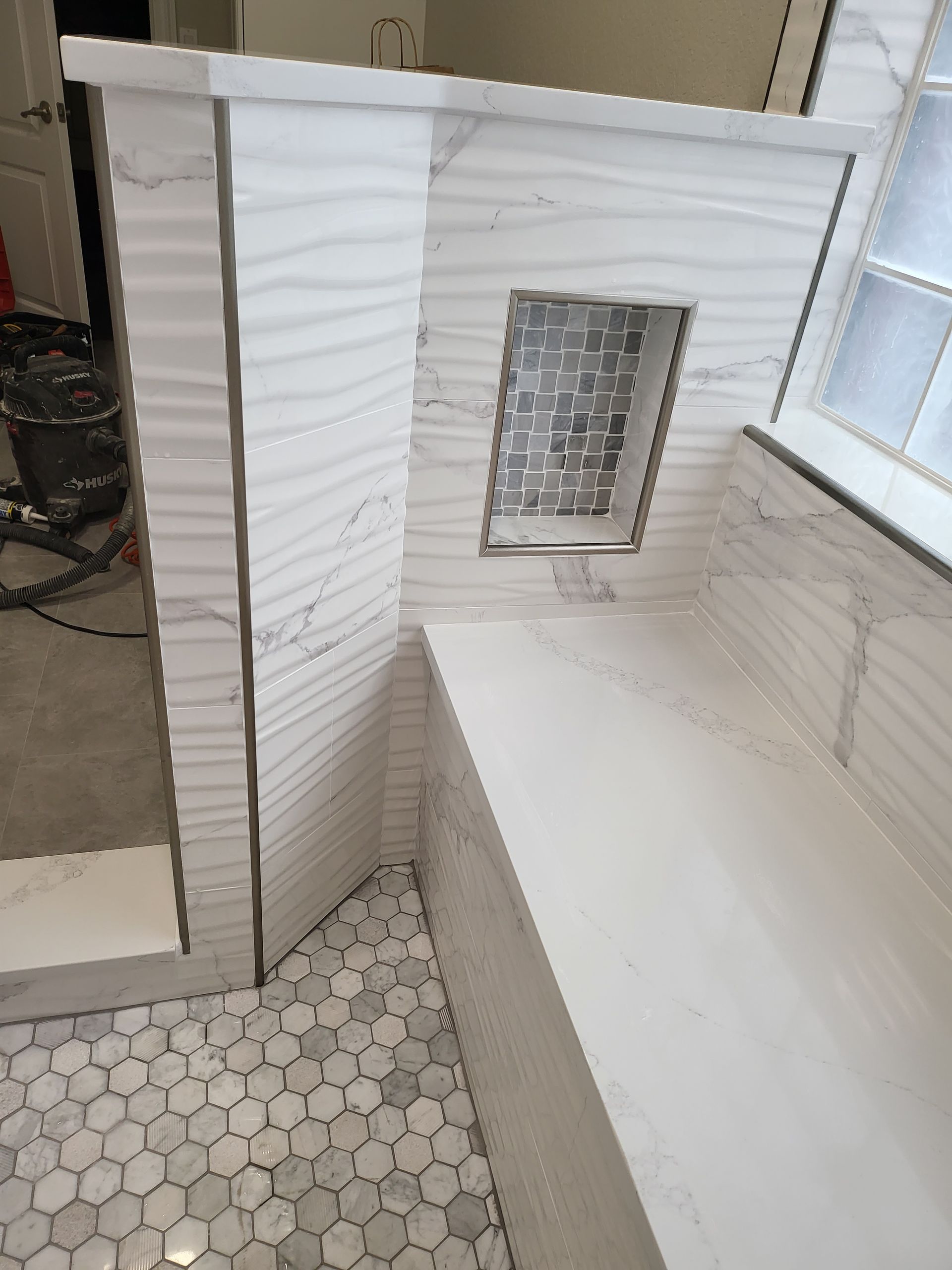 White tiled shower corner with built-in bench, hex floor tiles, and a small window.