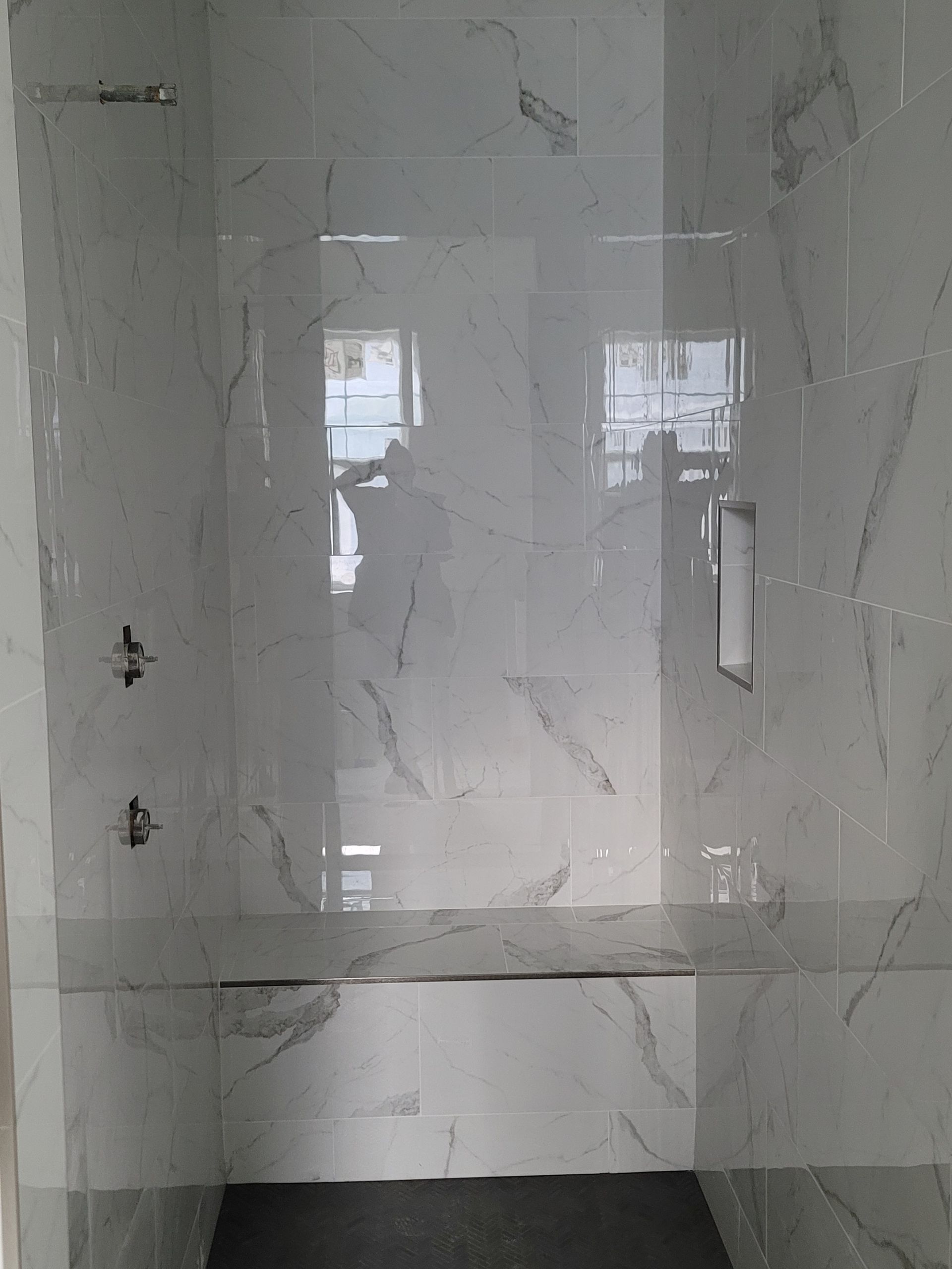 Empty tiled shower stall with glass door and chrome fixtures, reflecting two windows.