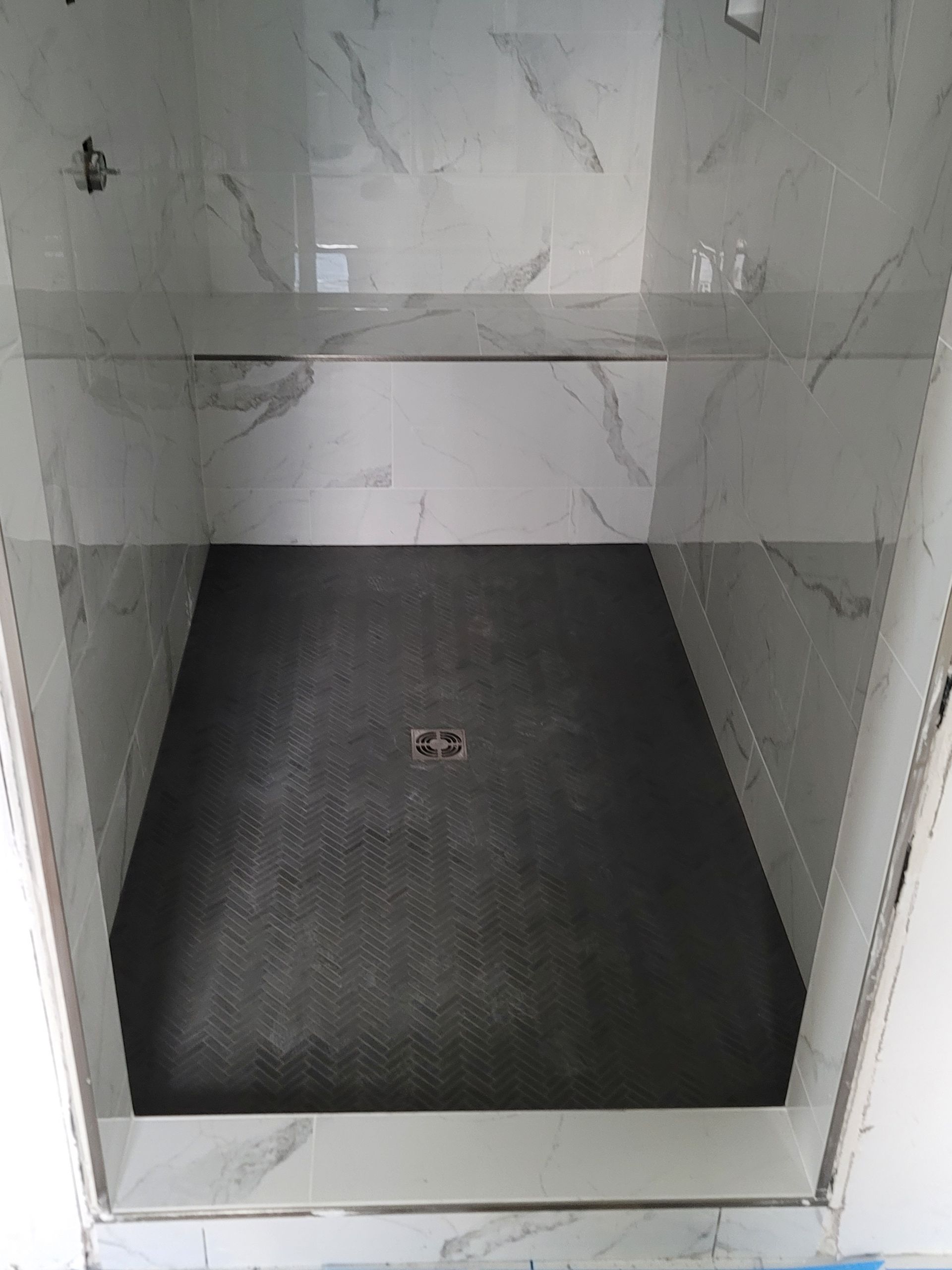 Empty tiled shower stall with a dark floor drain mat and light marble-pattern walls.