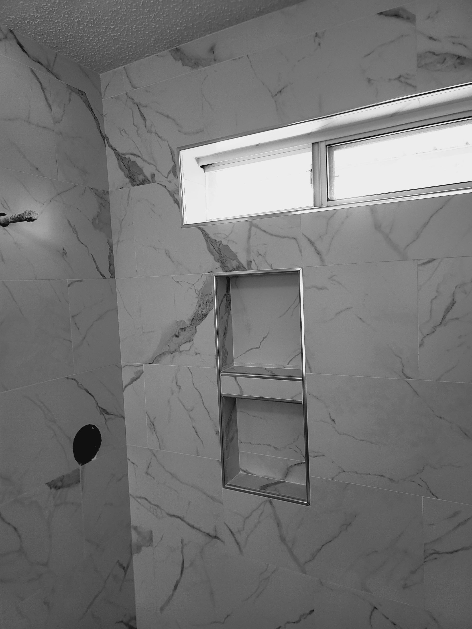 Tiled bathroom wall with small window and recessed shelves, unfinished cutout visible.