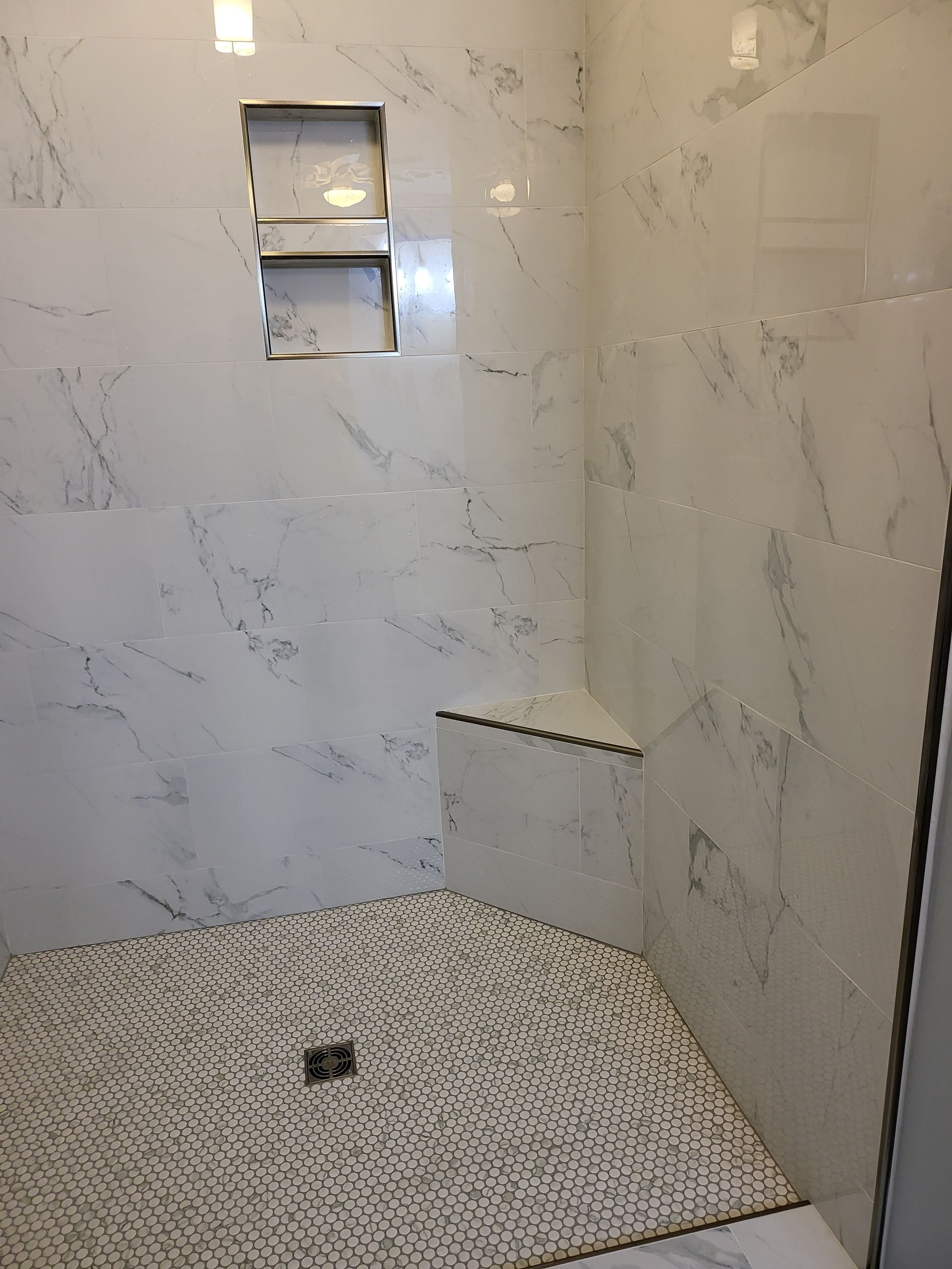 Empty tiled shower stall with marble-look walls, built-in shelves, and a pebble-pattern floor.