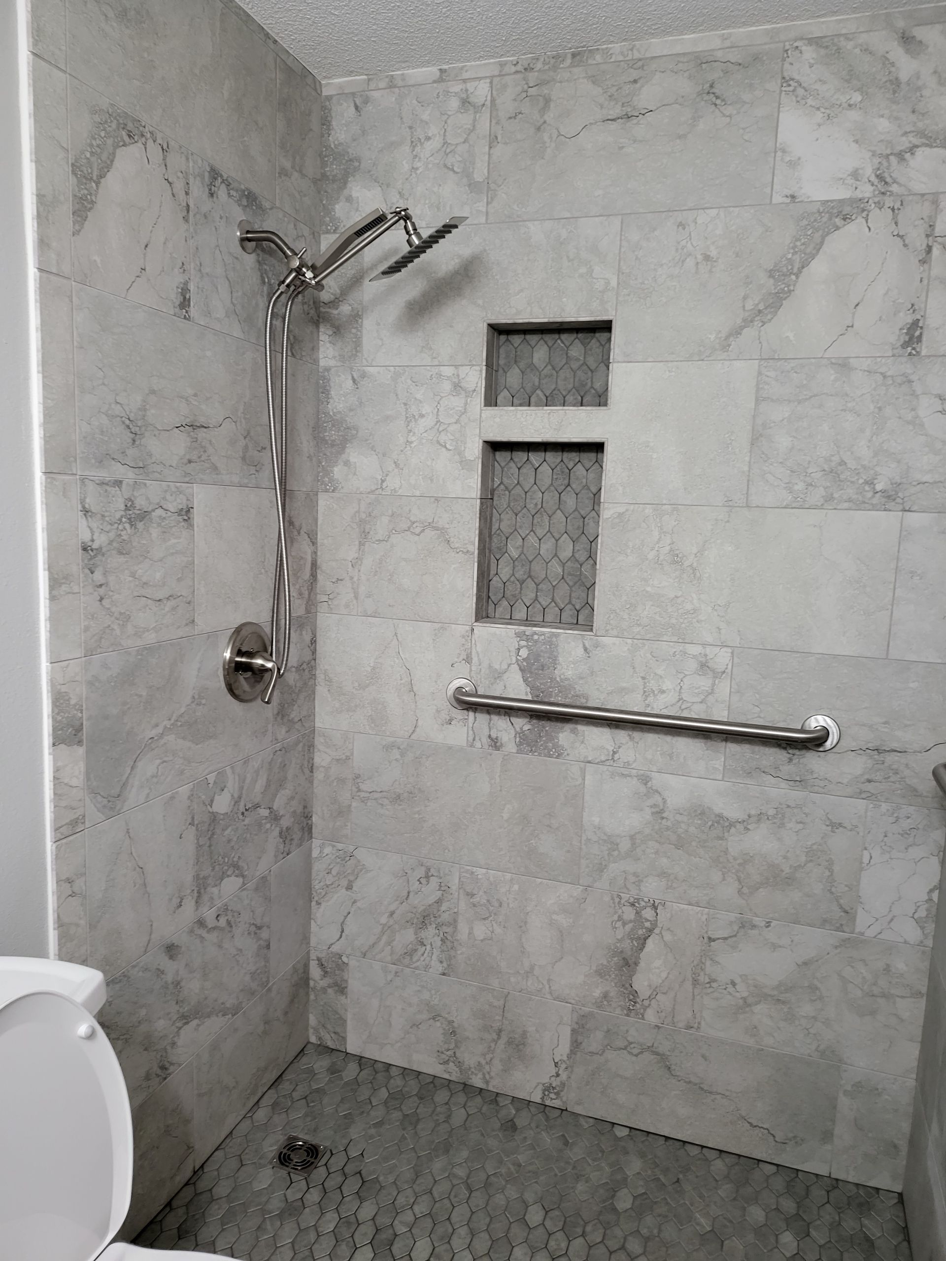 Accessible shower with marble tile walls, handheld showerhead, bench, and grab bar, viewed from the toilet area
