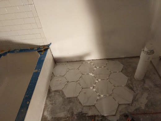 Bathroom corner with hexagonal tile floor, unfinished grout, and a blue-taped edge near the tub
