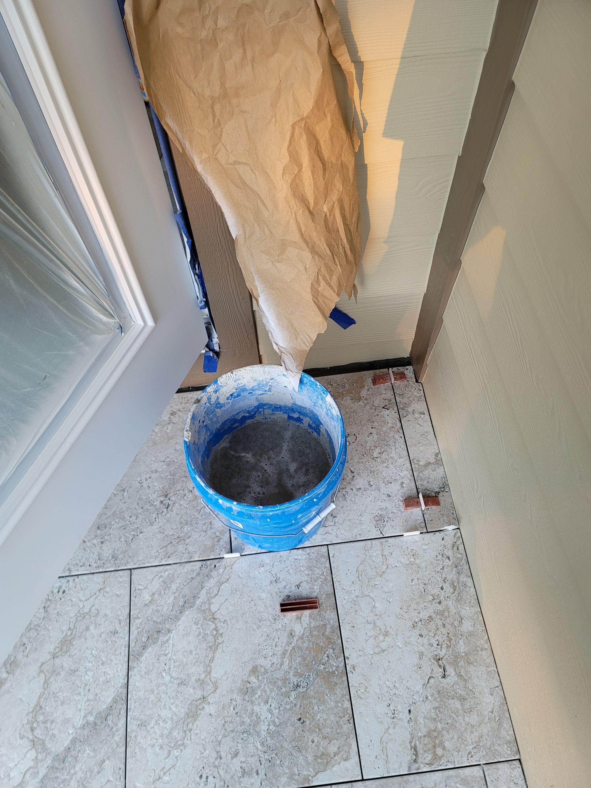 Blue bucket on tiled floor beside a window and draped paper in a narrow indoor corner.