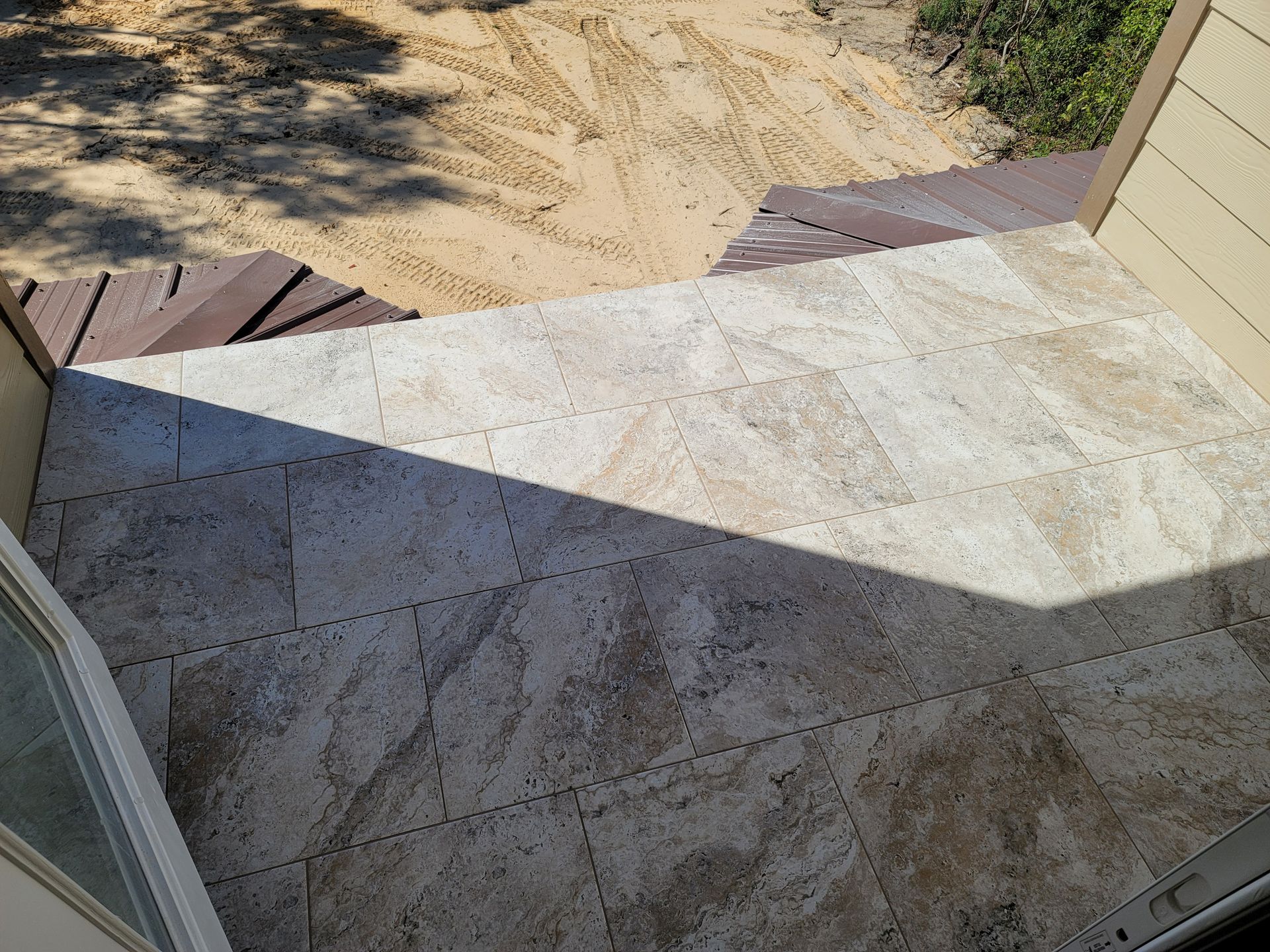 Stone patio and sandy yard beside a house, with sharp sunlight and shadows across the walkway
