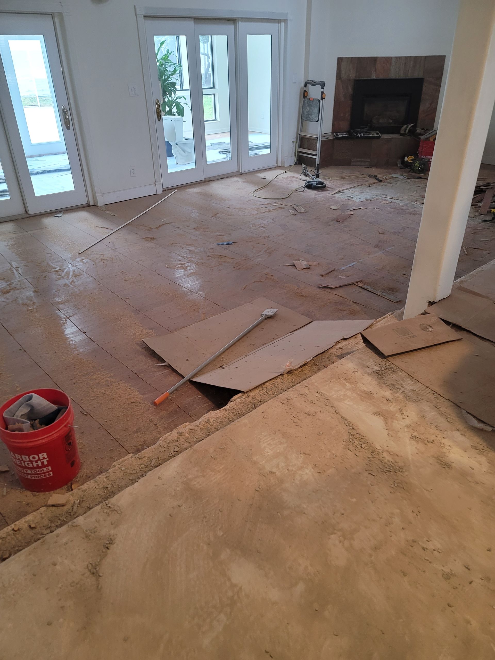 Living room under renovation with exposed flooring, scattered debris, and a fireplace in the back.